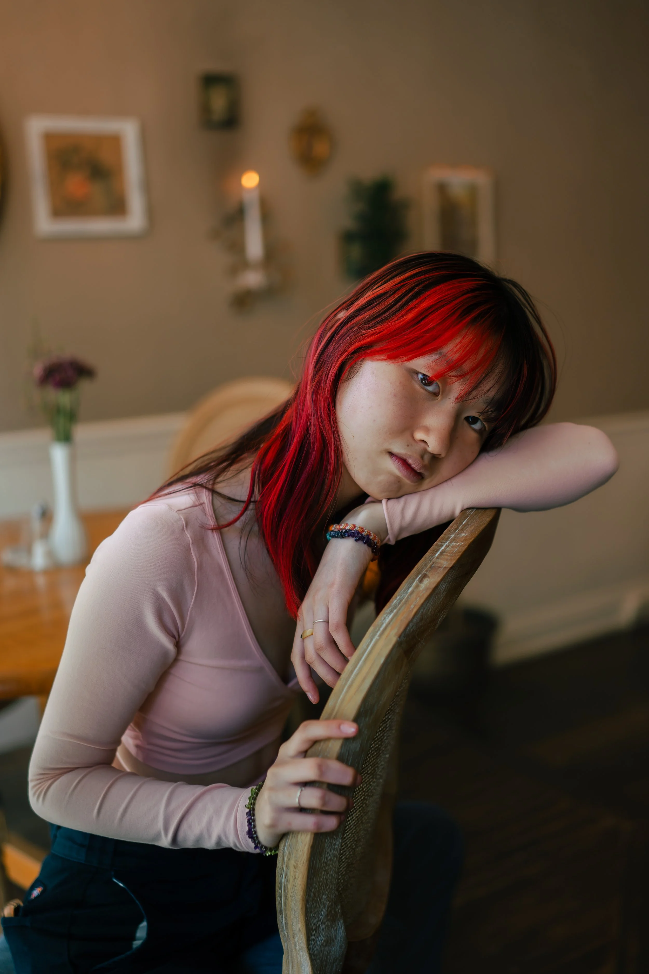 A young woman with red highlighted hair leaning on a wooden chair in a cozy, decorated room.