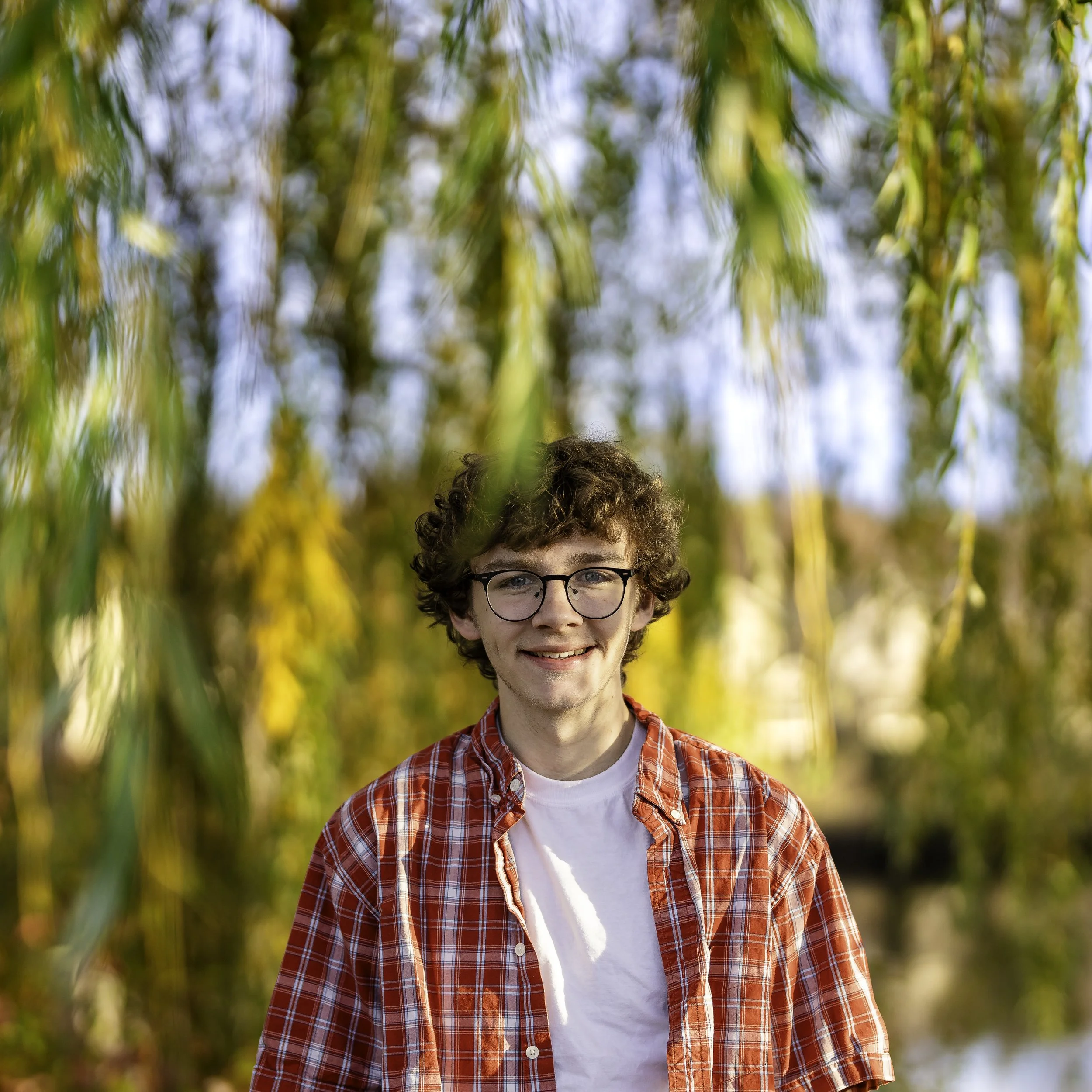 A young man with curly brown hair and glasses smiling outdoors among trees with yellow and green leaves in the fall.