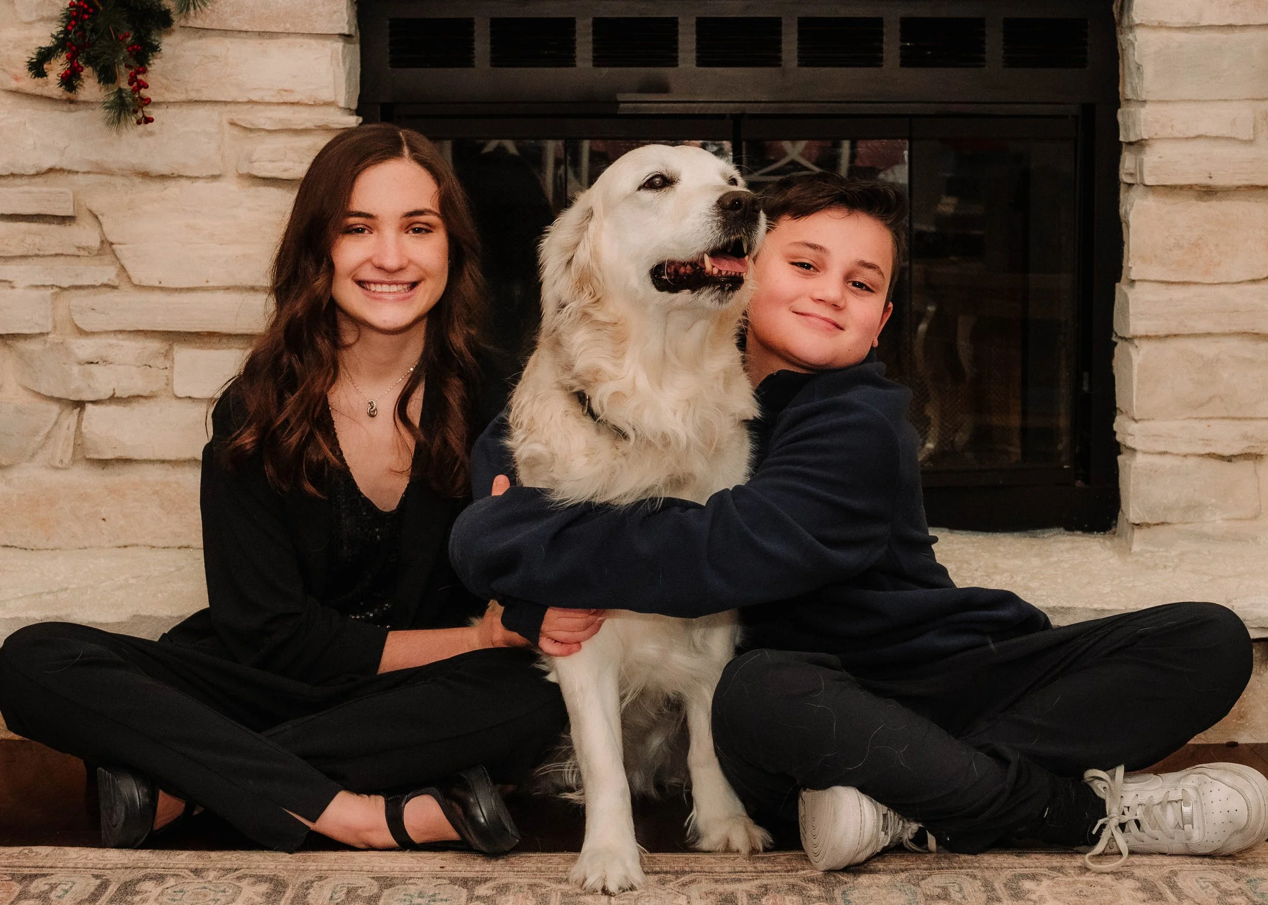 Two children, a girl and a boy, sitting on the floor in front of a fireplace with their golden retriever dog, all smiling and embracing.
