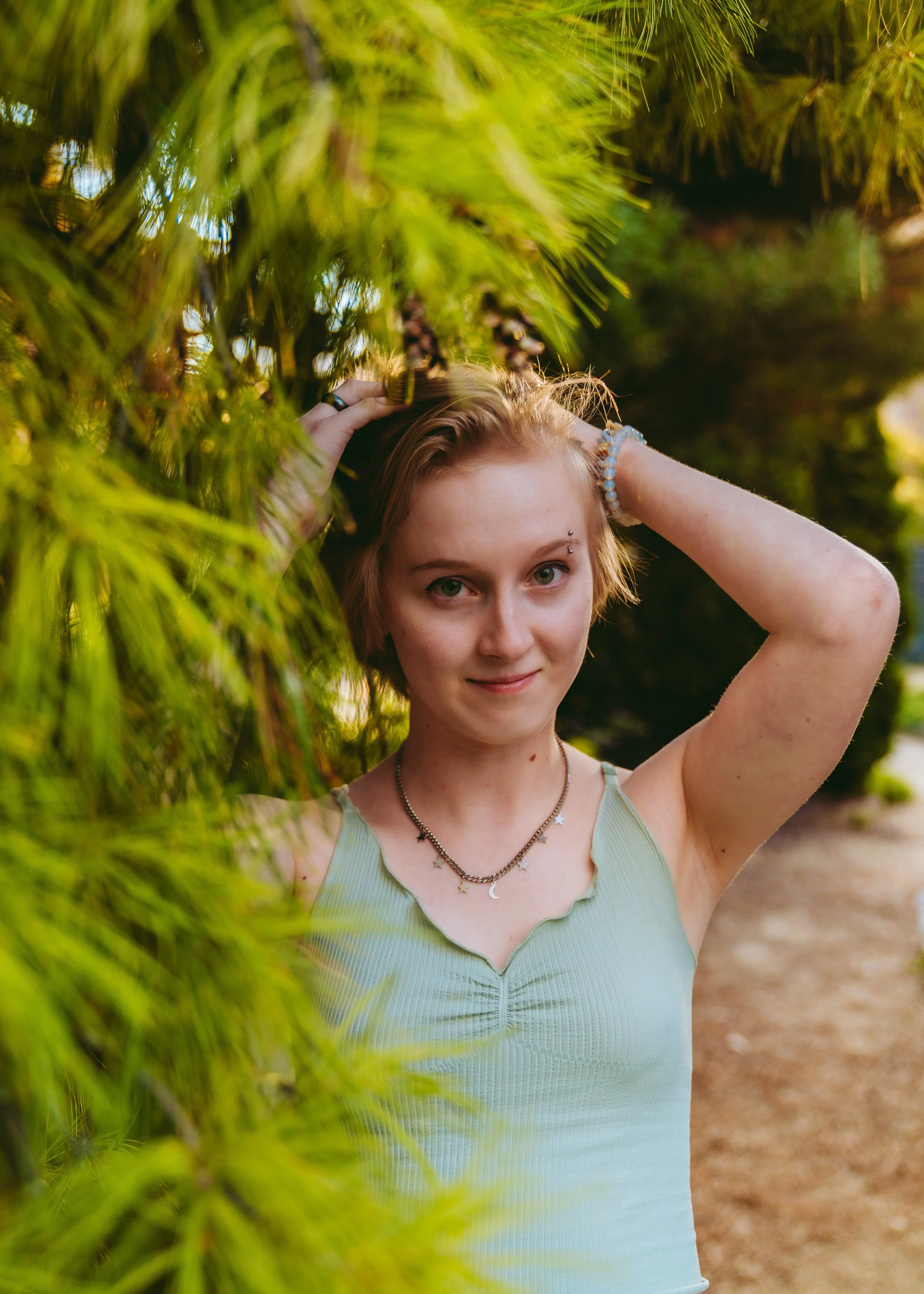 A young woman with short brown hair and piercings on her eyebrow and nose, wearing a light green sleeveless top, stands outdoors among green pine trees. She is smiling and has her hands raised to her head, with one hand resting on her hair and the ot