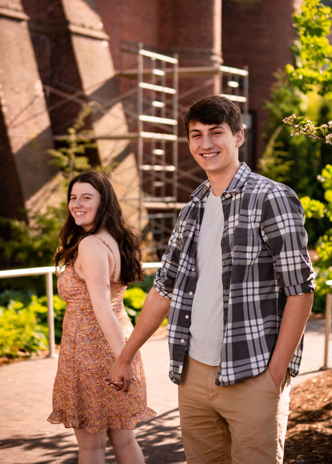 A young couple holding hands and smiling outside on a sunny day, with a brick building and greenery in the background.