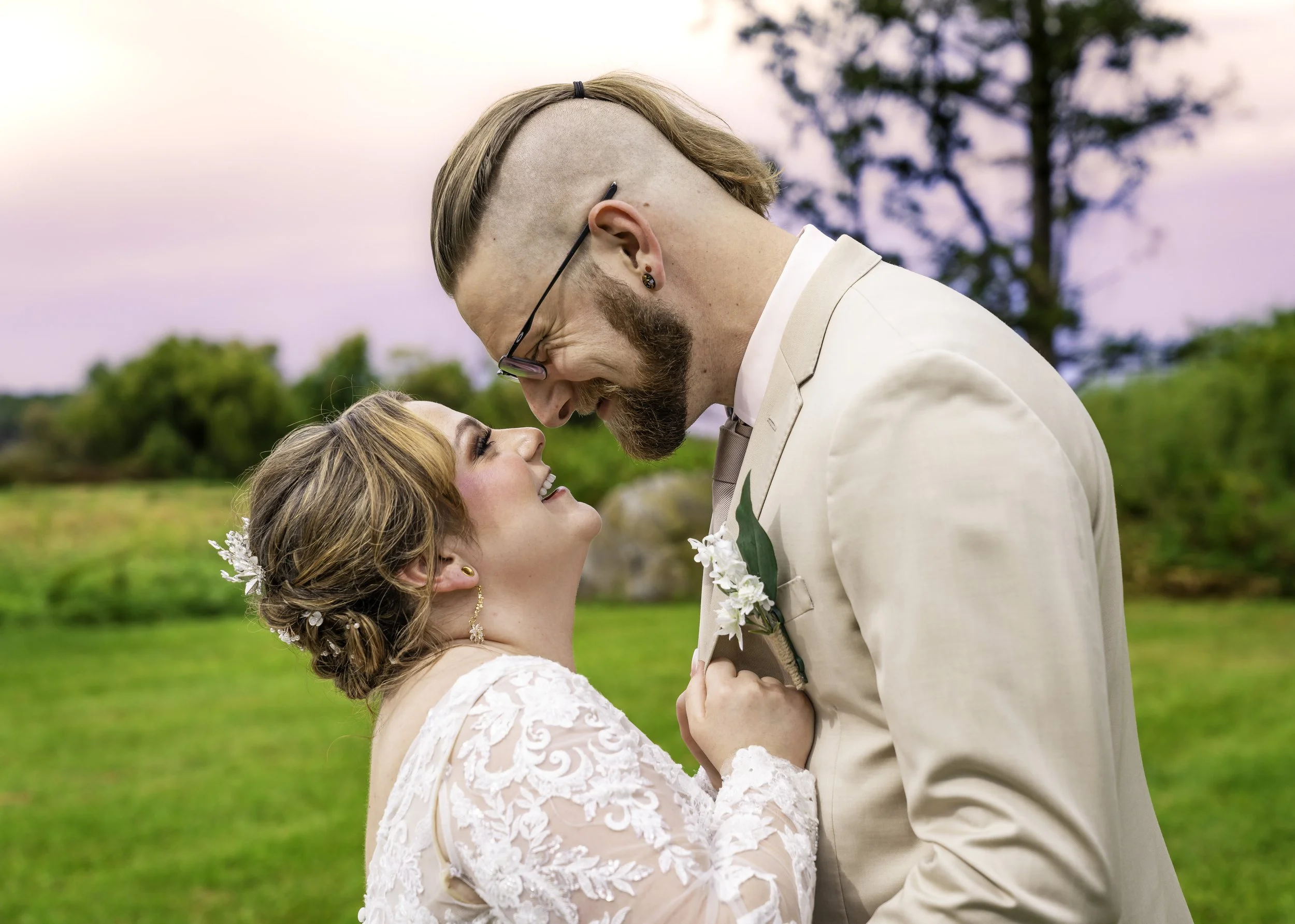 A bride and groom smiling and looking into each other's eyes outdoors during their wedding, with green trees and grass in the background.