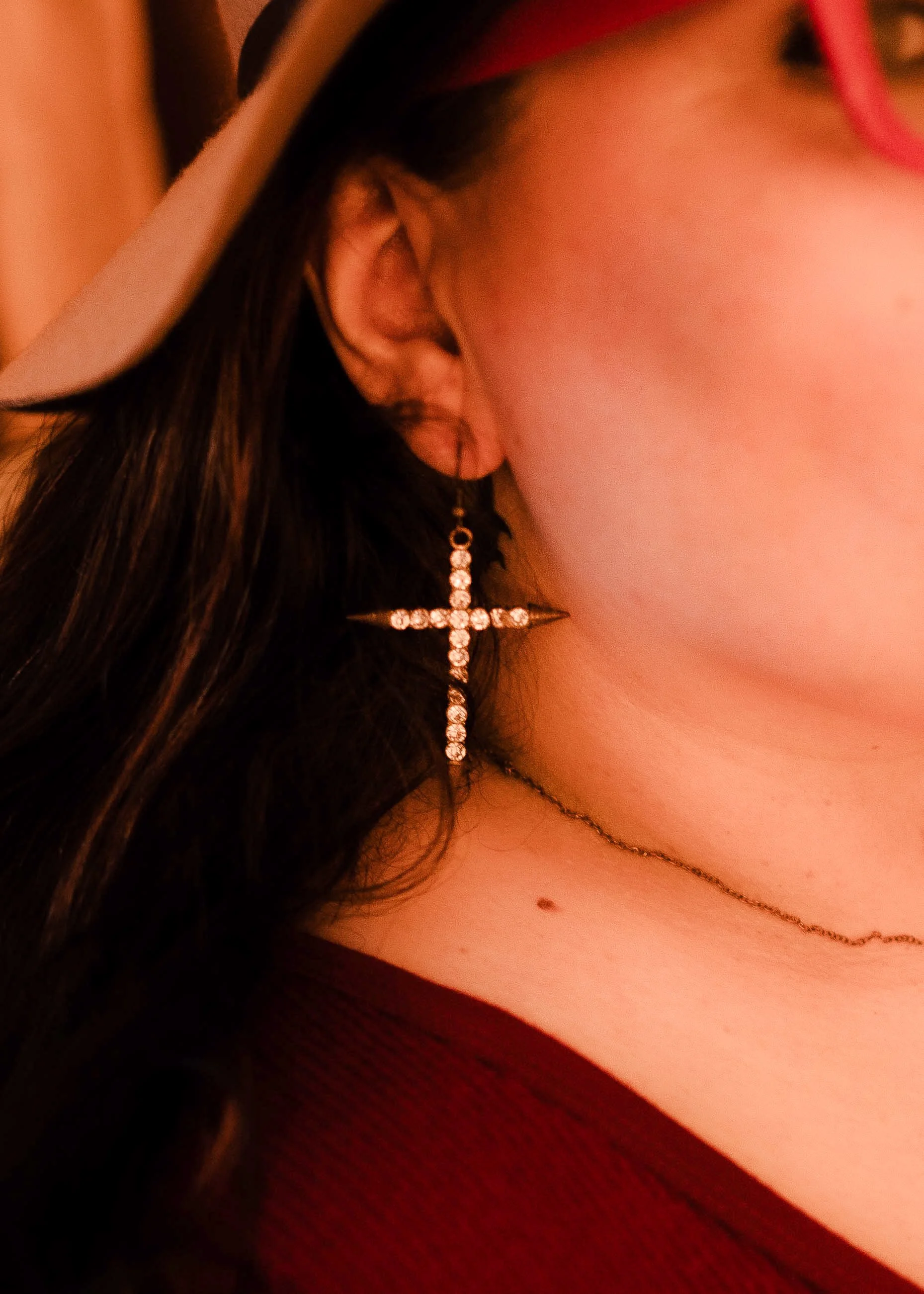 Close-up of a woman wearing a large cross-shaped earring with rhinestones, a thin chain necklace, and pink glasses, with a portion of her face visible.