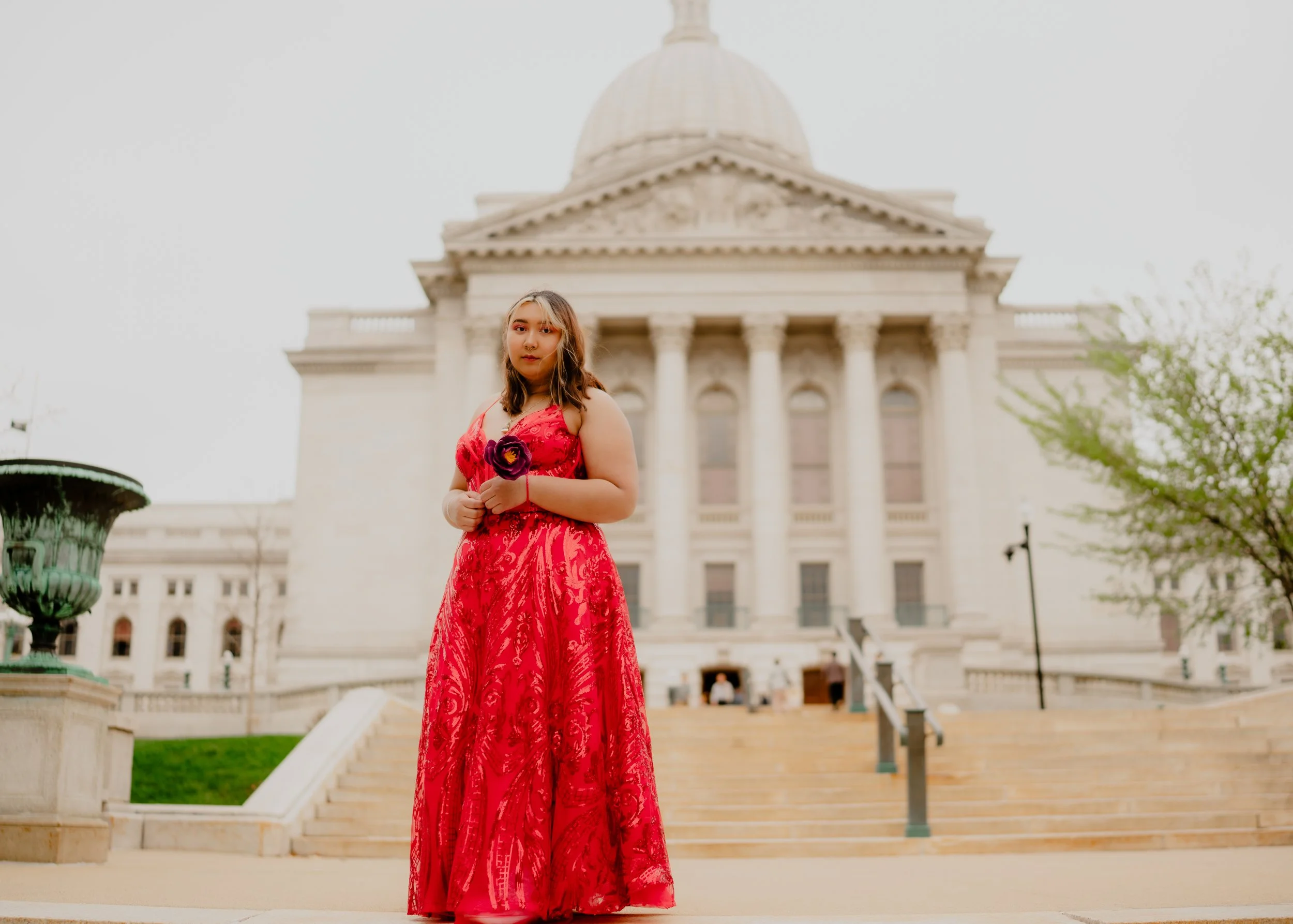 Young woman in a red dress holding a purple flower standing in front of a large government or historic building with steps and columns.