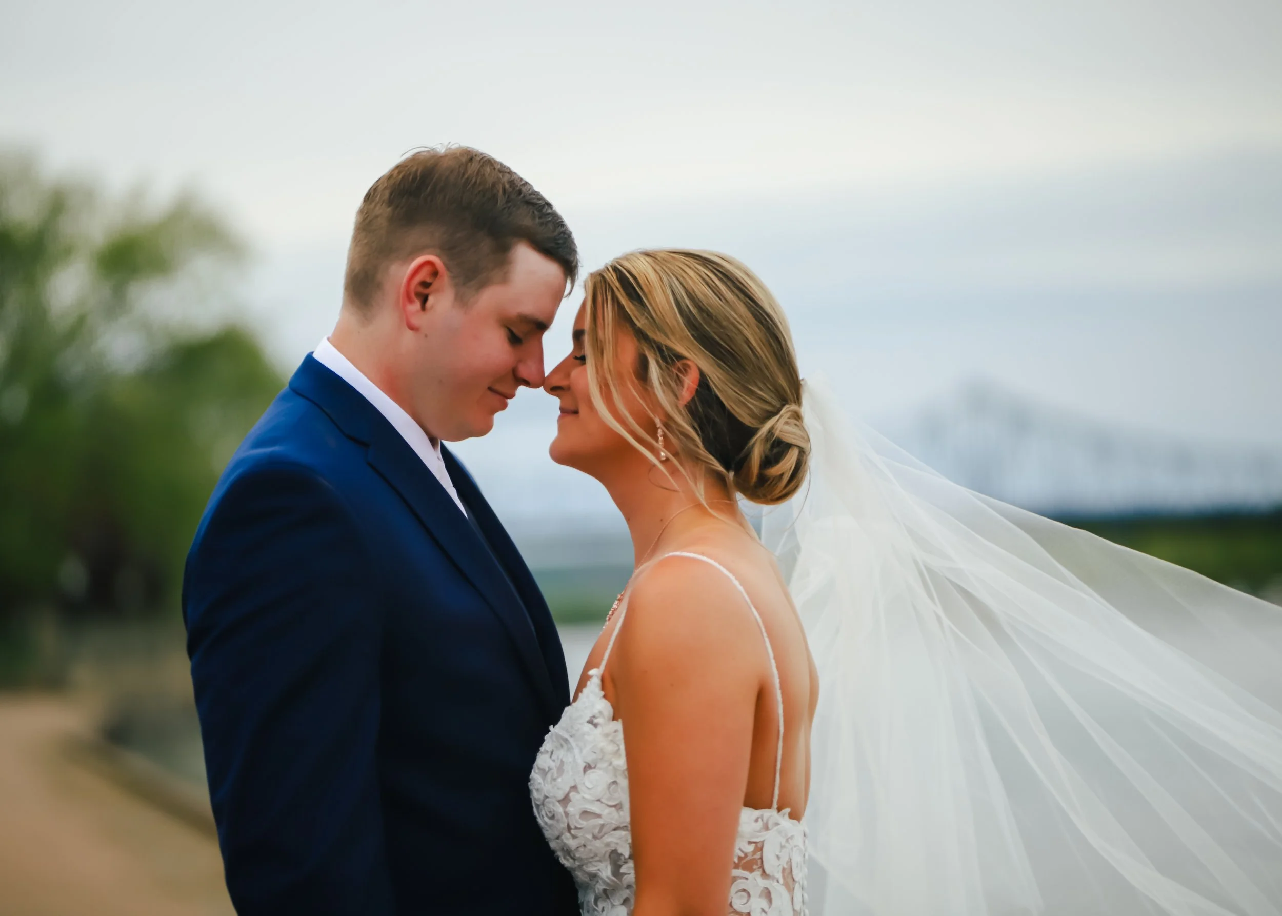 A bride and groom with foreheads touching, smiling, outdoors, wedding attire, overcast sky, with a blurred bridge in the background.