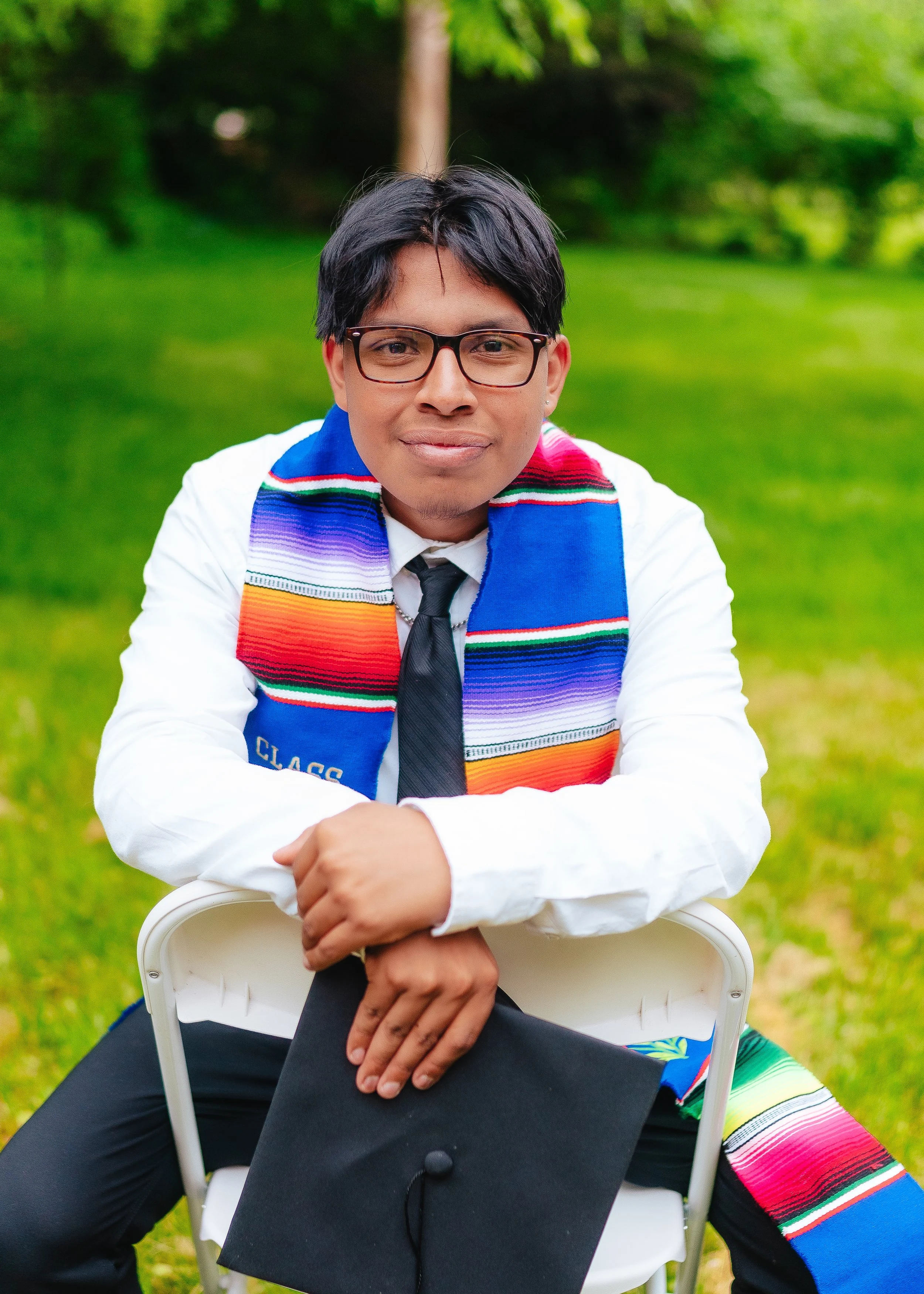 Young man in a white shirt, black tie, and glasses sitting outdoors with a colorful scarf, holding a graduation cap, in a green park.