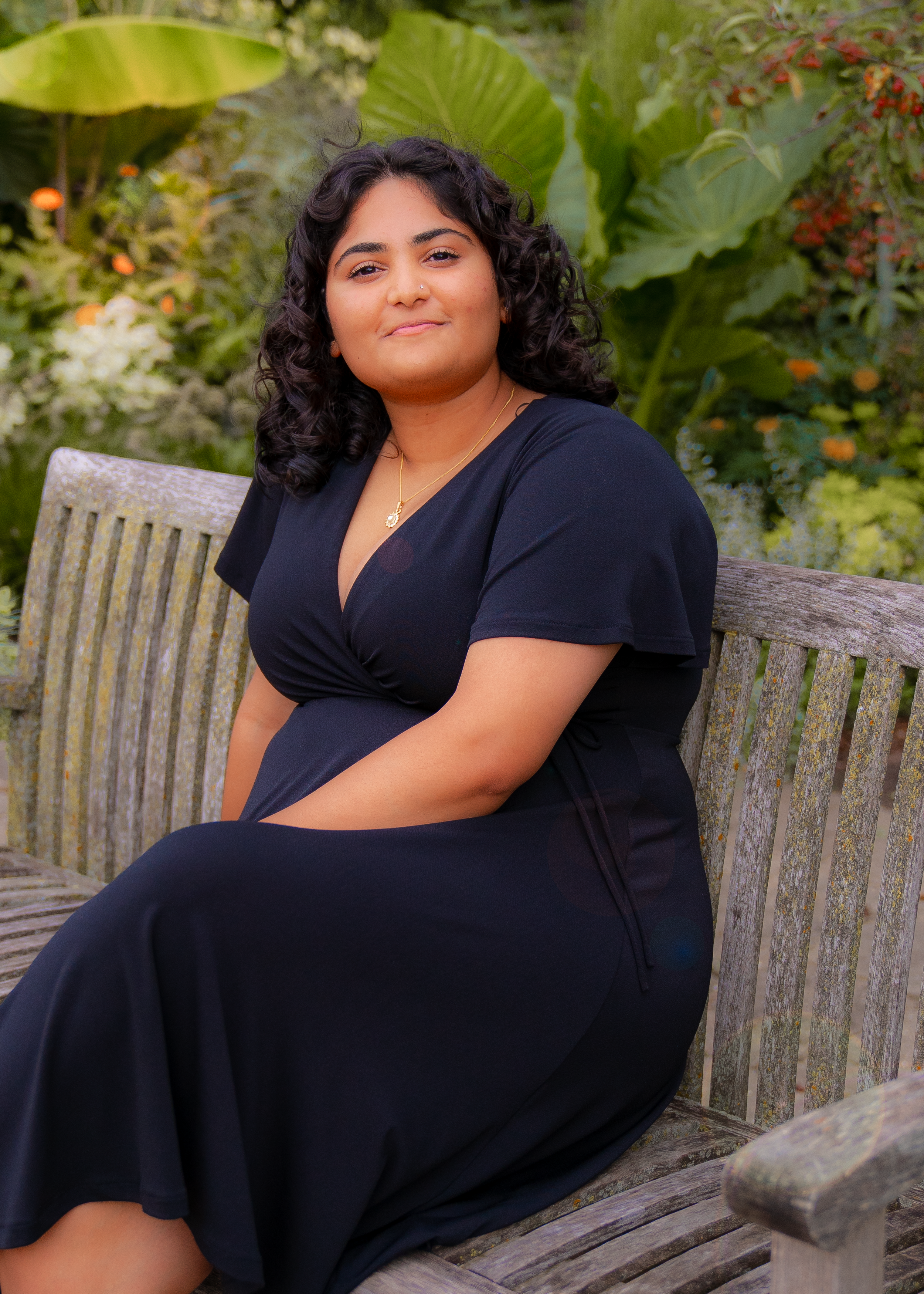 A woman with dark curly hair sitting on a weathered wooden bench in a lush garden with large green leaves and flowers around her, wearing a black dress and a gold necklace.