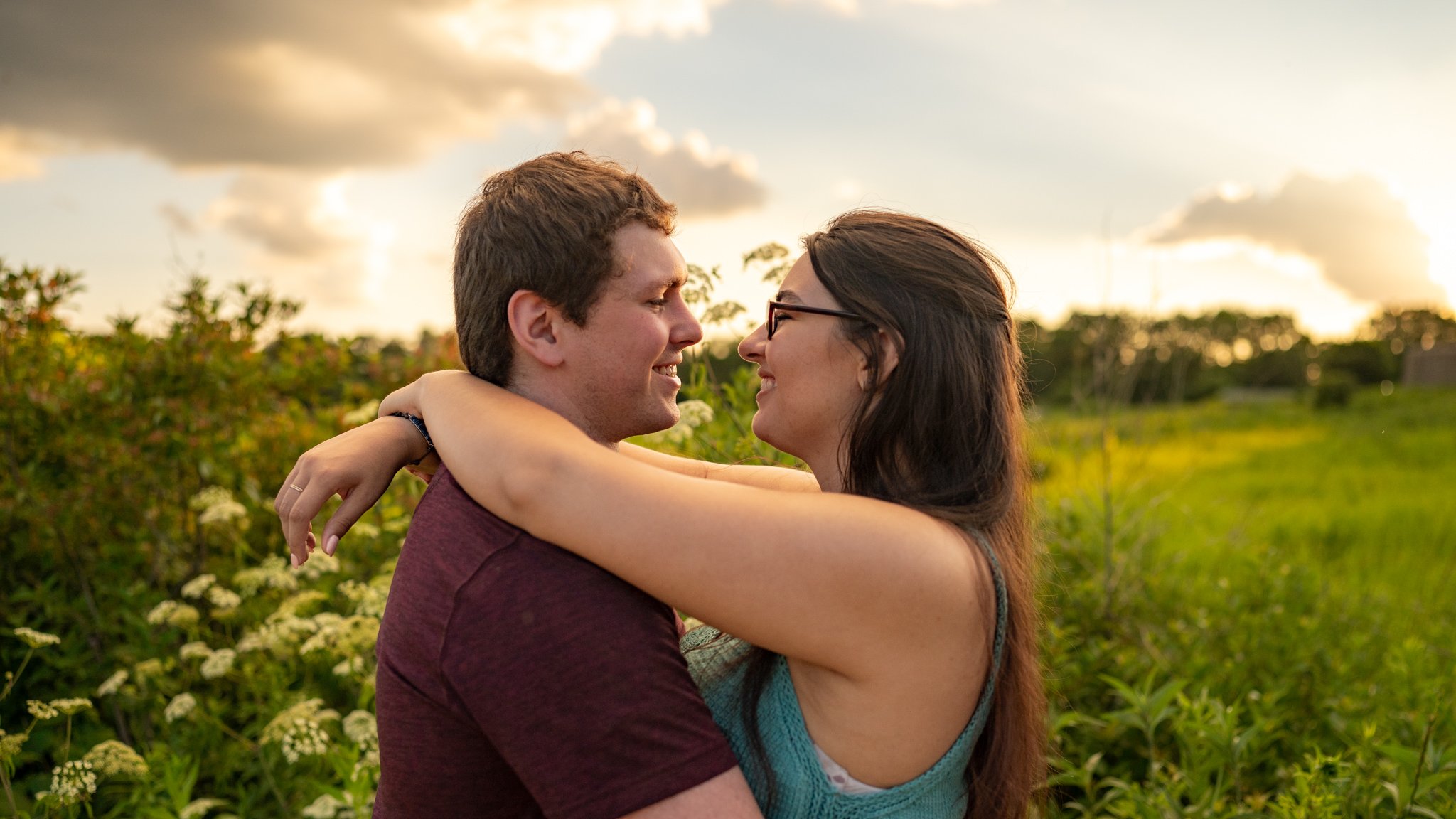 A young couple embracing outdoors in a field during sunset, smiling and facing each other.