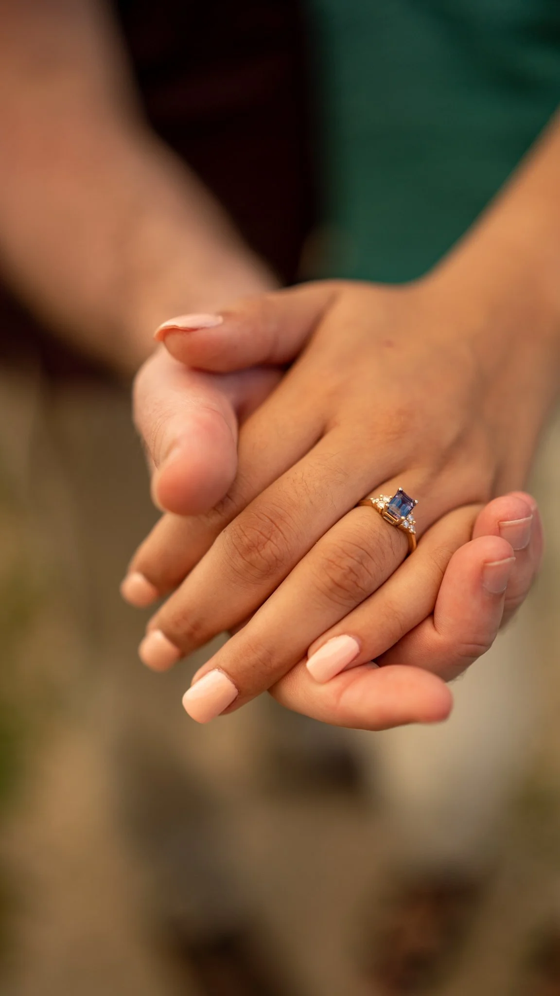 Close-up of a woman’s hand wearing an engagement ring with a blue gemstone and smaller diamonds, gently held by another person’s hand.