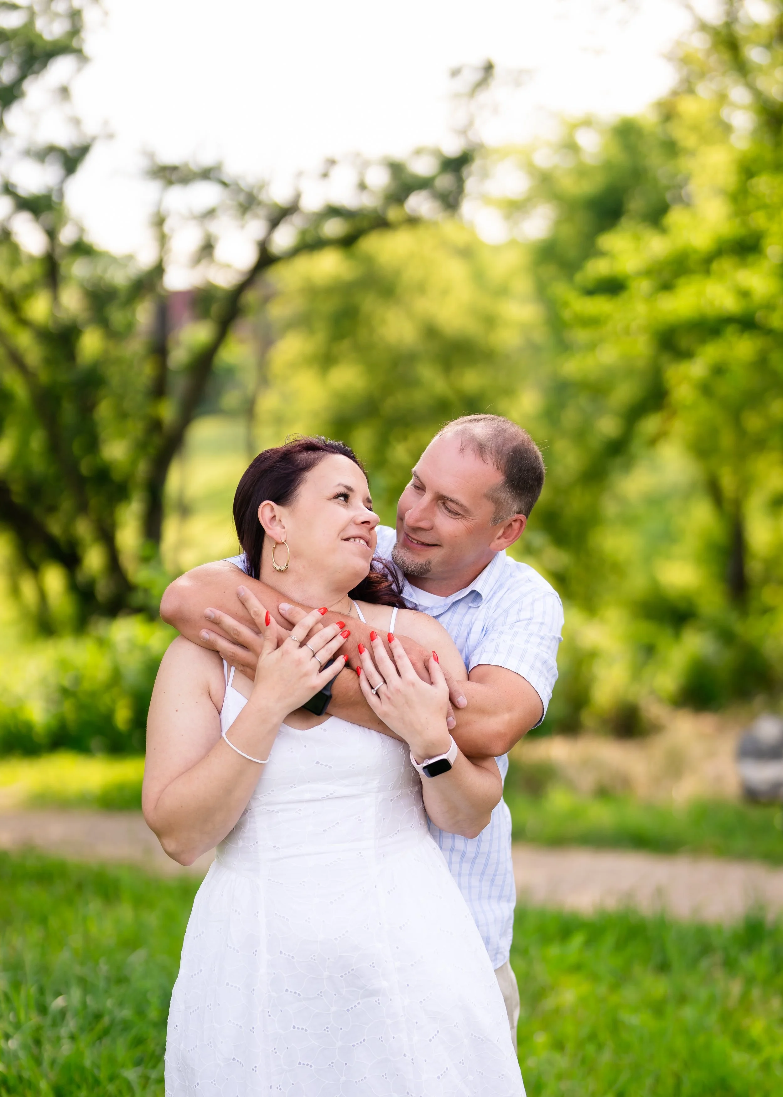 A couple embracing outdoors in a green park, smiling at each other.