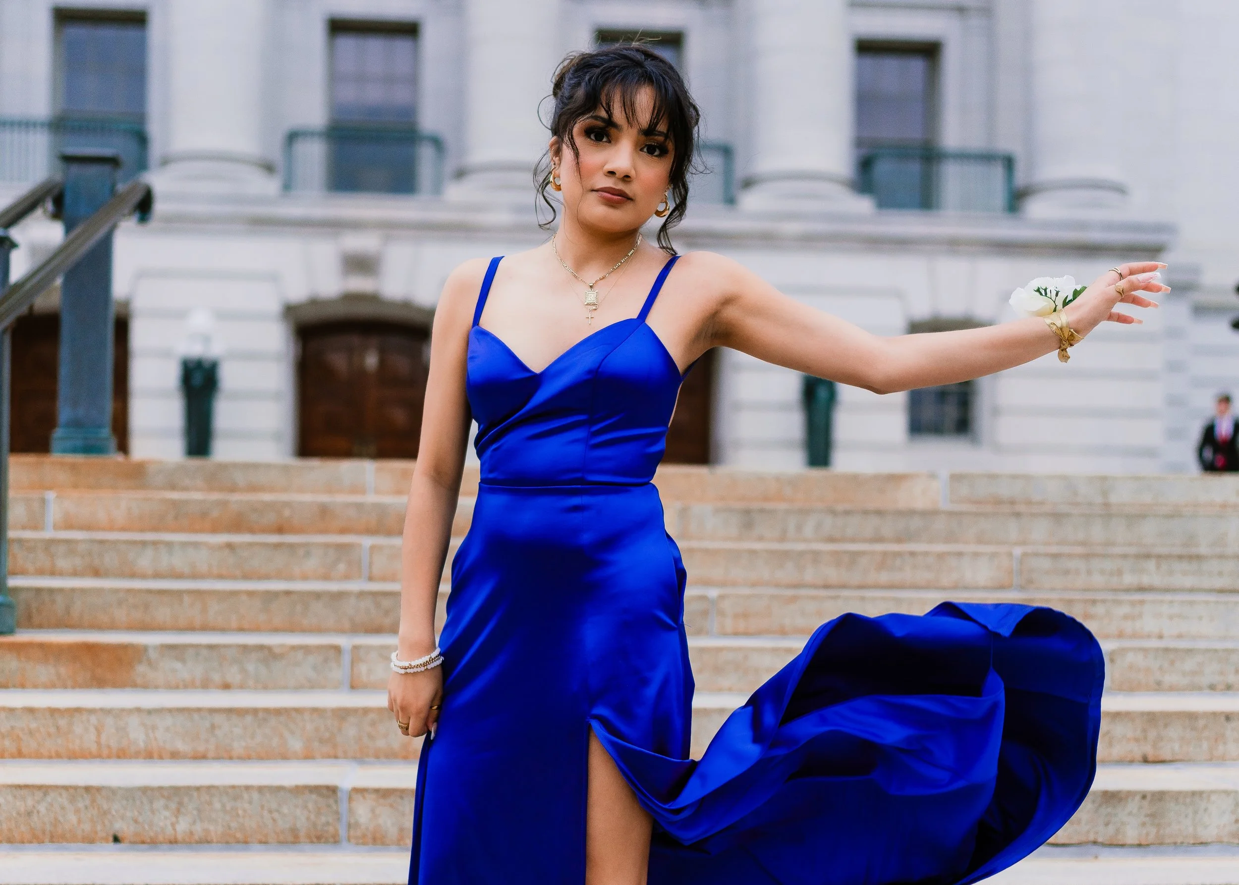 A woman in a vibrant blue gown with a high slit, posing on steps in front of a large classical-style building.