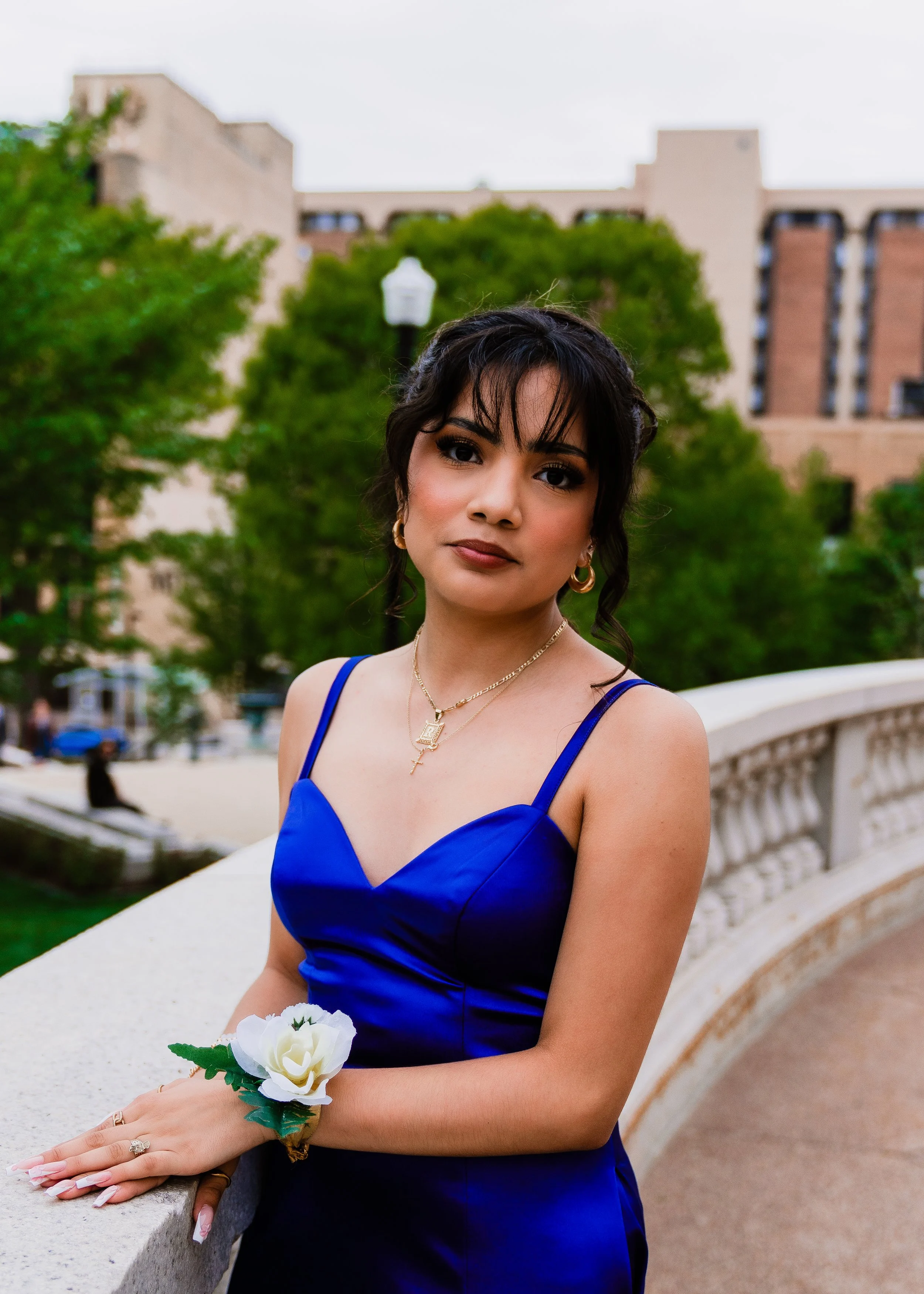A woman in a royal blue dress standing outdoors on a bridge, holding a white flower bracelet, with green trees and a building in the background.