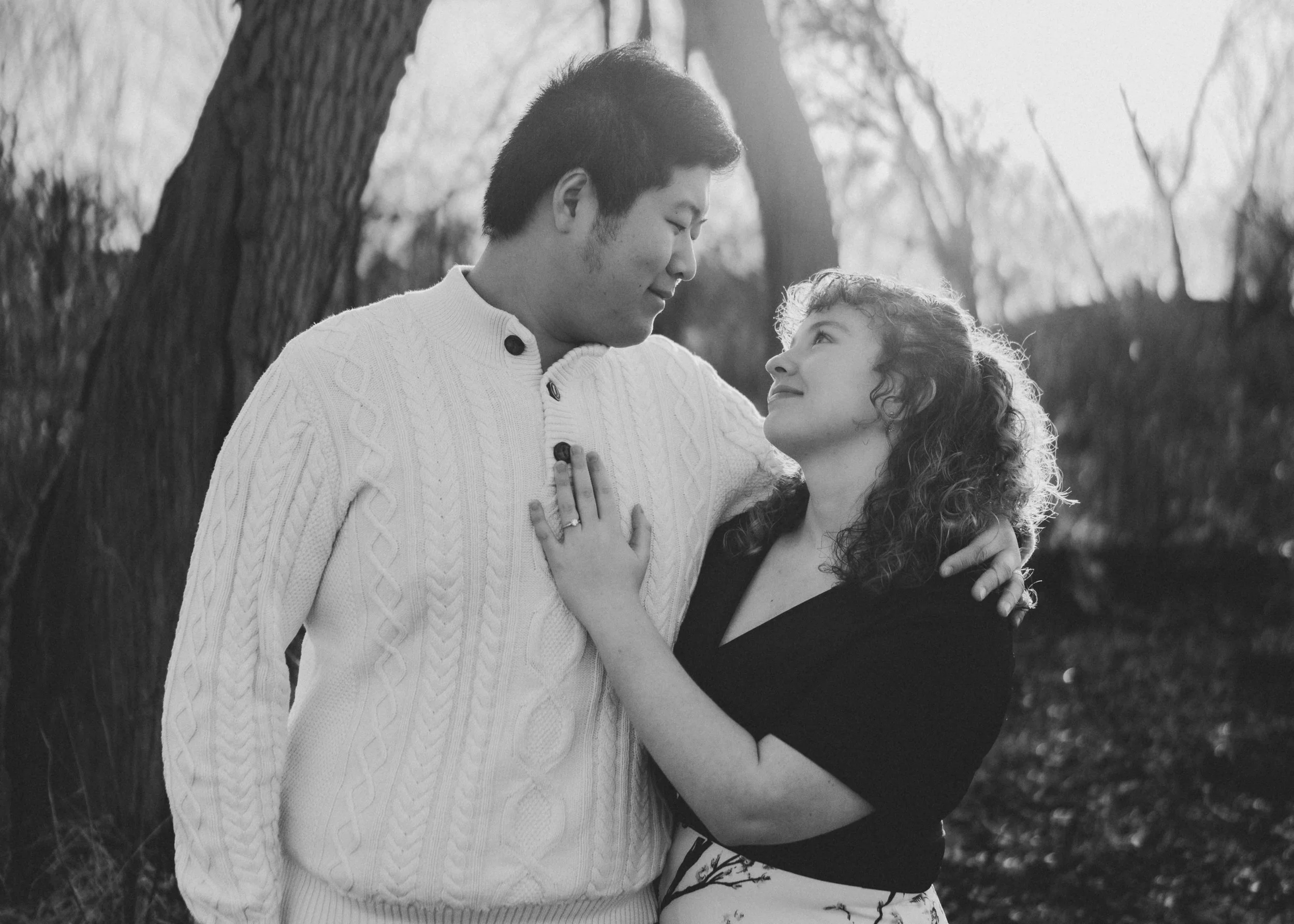 A man and woman stand close together outdoors in a black-and-white photo, gazing into each other's eyes. The man is wearing a white cable-knit sweater, and the woman is wearing a black top with curly hair. The background includes trees and sunlight f