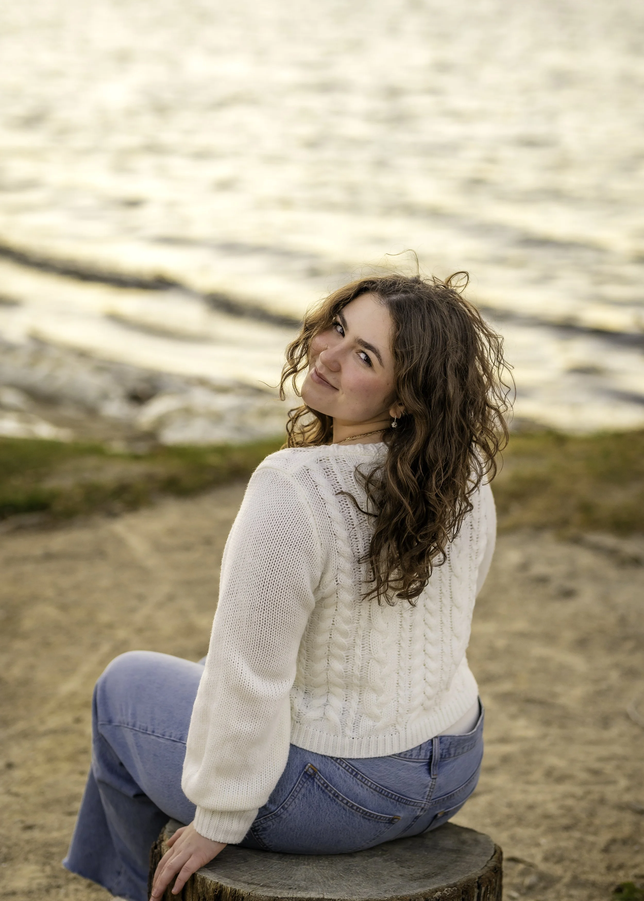 A woman with curly brown hair sitting on a tree stump near a body of water, smiling and looking back at the camera during sunset.
