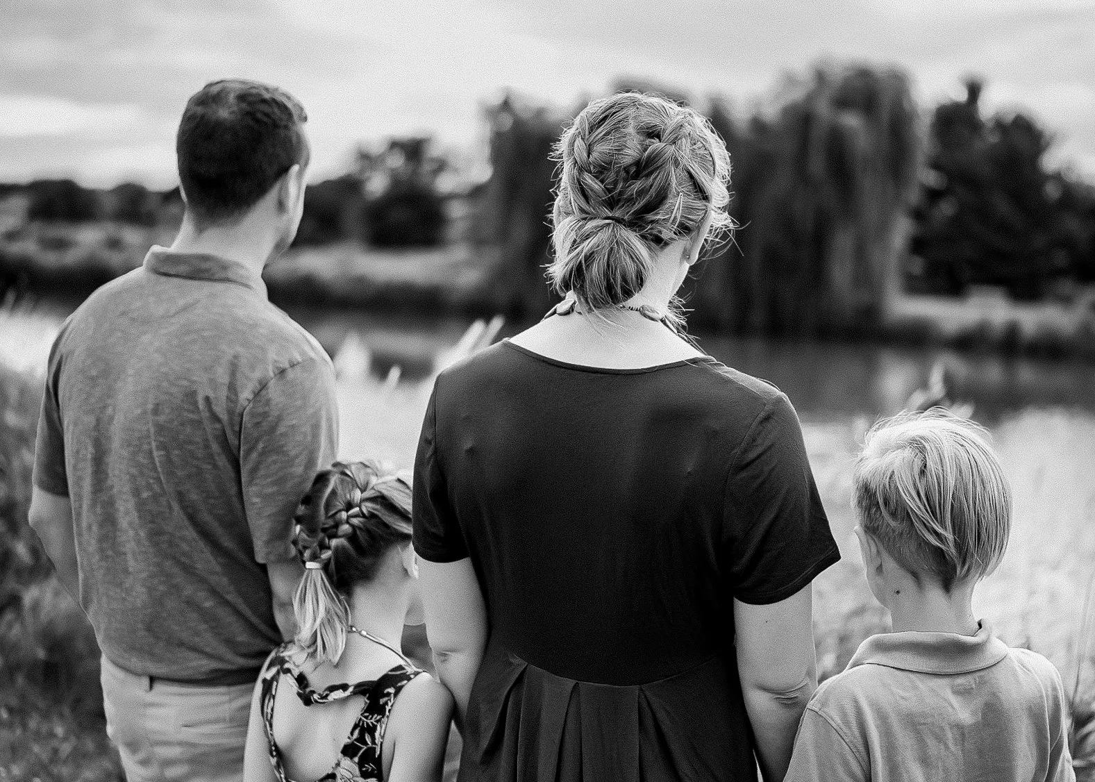 A black and white photo of four people, an adult woman and three children, standing outdoors near a body of water with trees in the background. They are looking away from the camera.