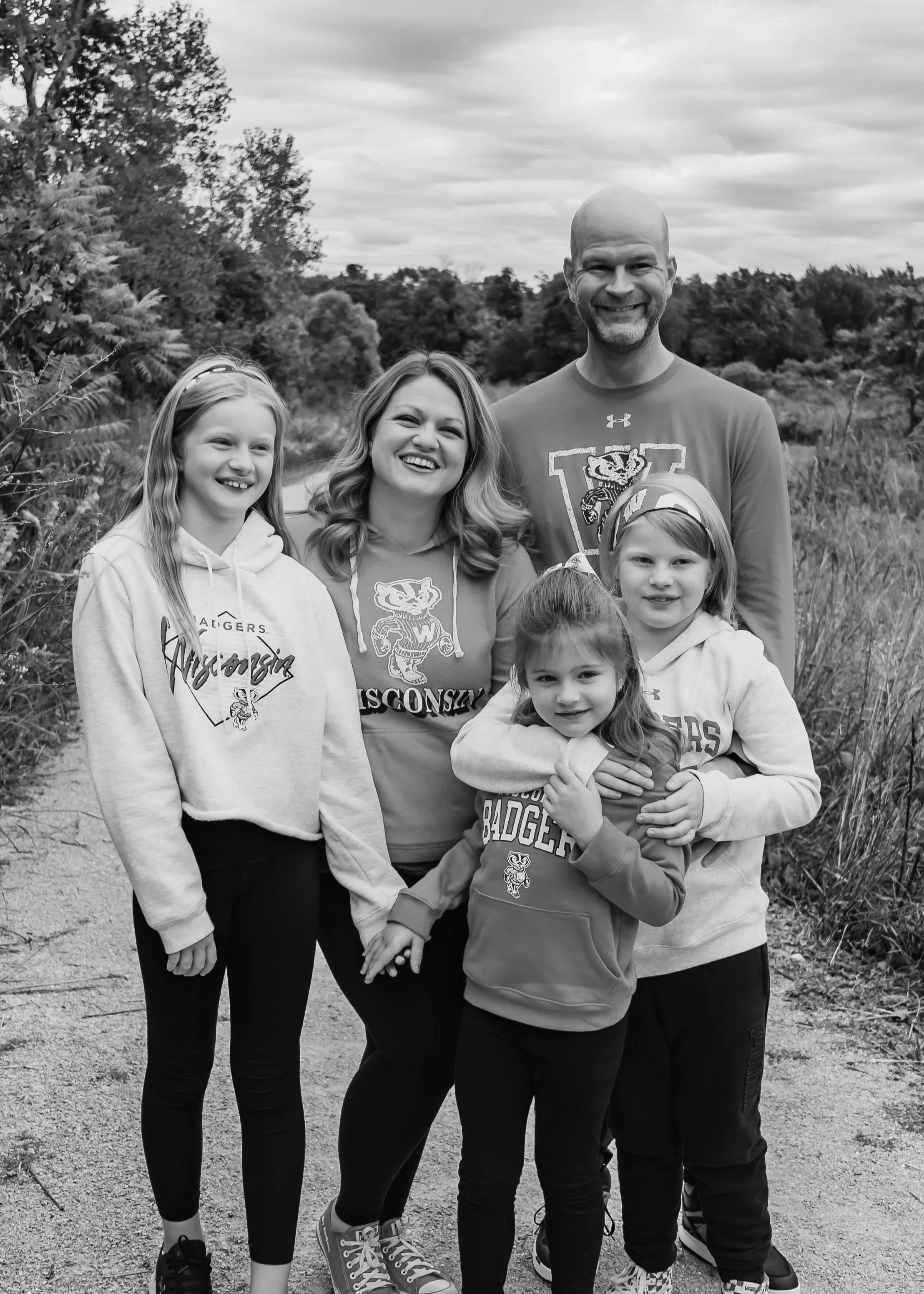 A family of six, including three children and two adults, standing outdoors on a nature trail, wearing Wisconsin Badgers clothing, smiling, with trees and cloudy sky in the background.
