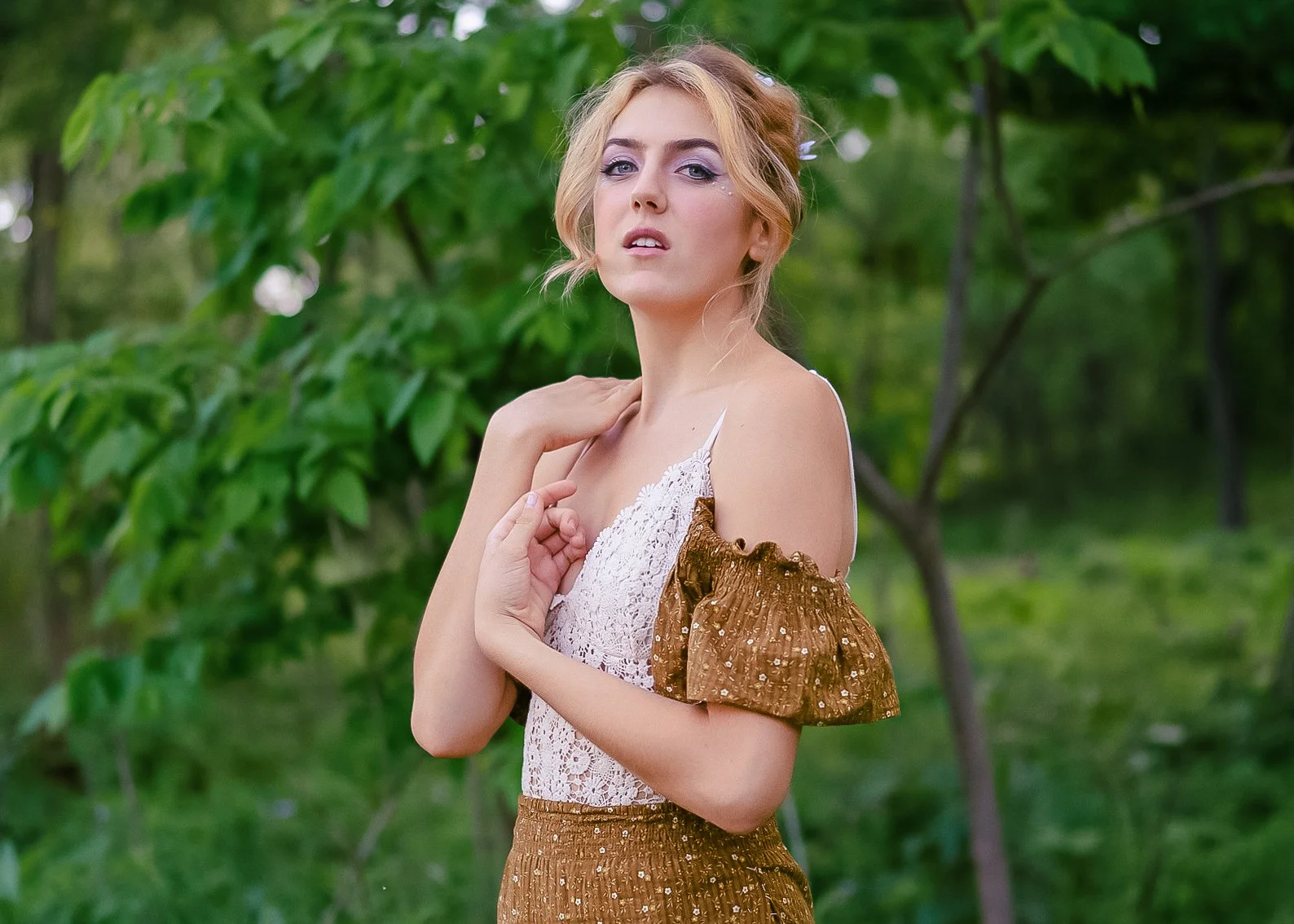 A woman with blonde hair and purple eyeshadow posing outdoors among green trees, wearing a lace top with ruffled brown sleeves and matching brown skirt.