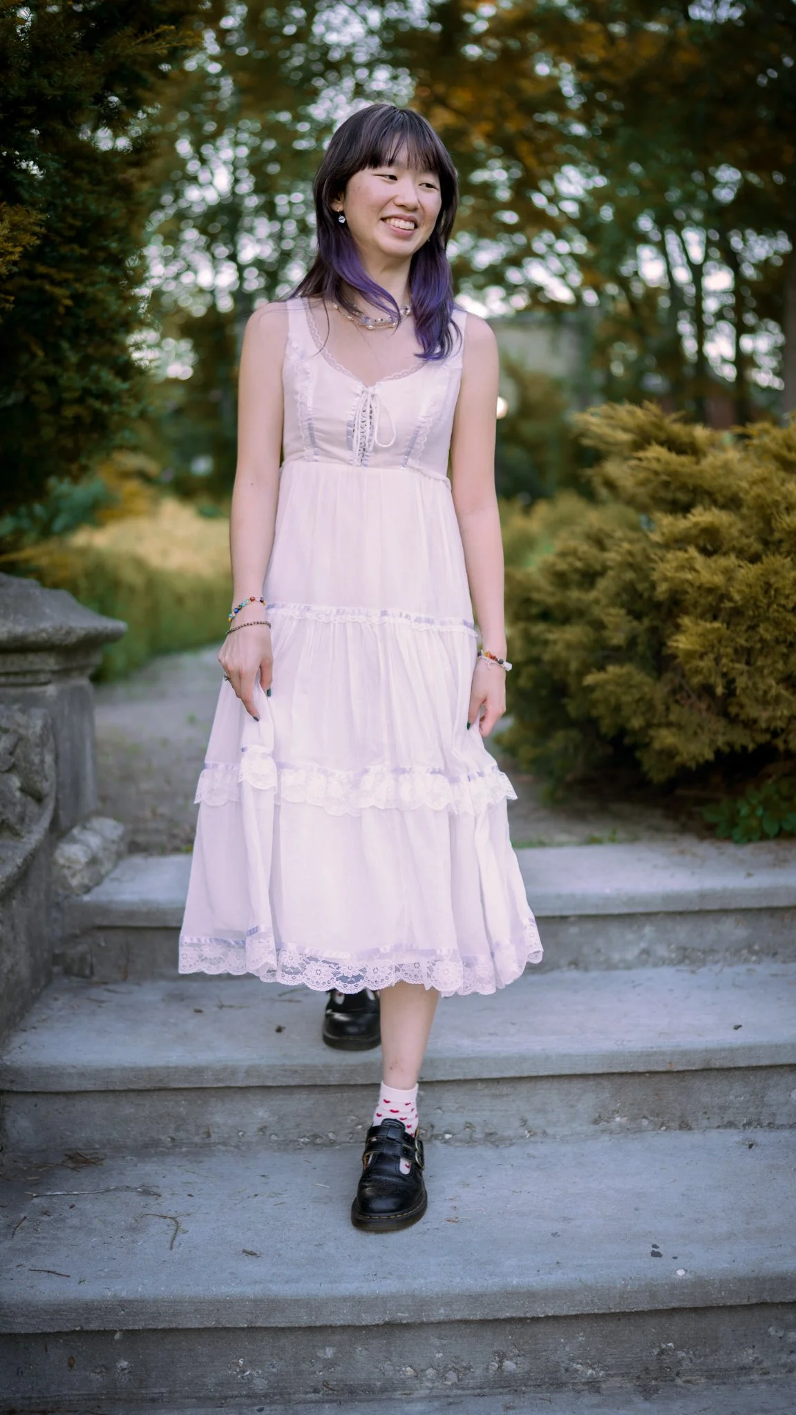A young woman in a white, sleeveless, tiered dress with lace trim is walking down concrete steps outside. She has dark hair with purple ends and is smiling. The background features greenery, trees, and bushes.
