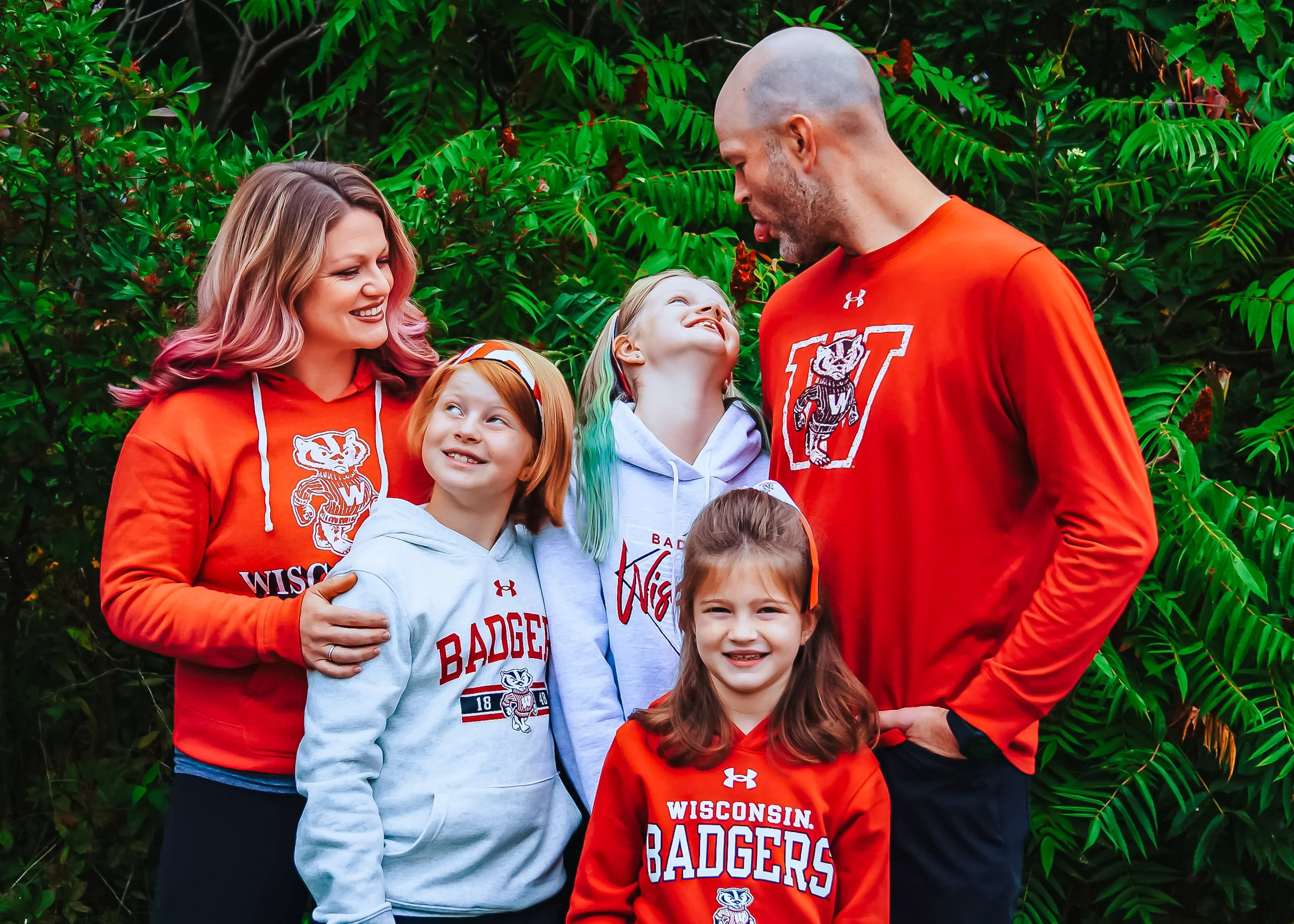 Family of six wearing Wisconsin Badgers apparel standing outdoors in front of green foliage, smiling and interacting.