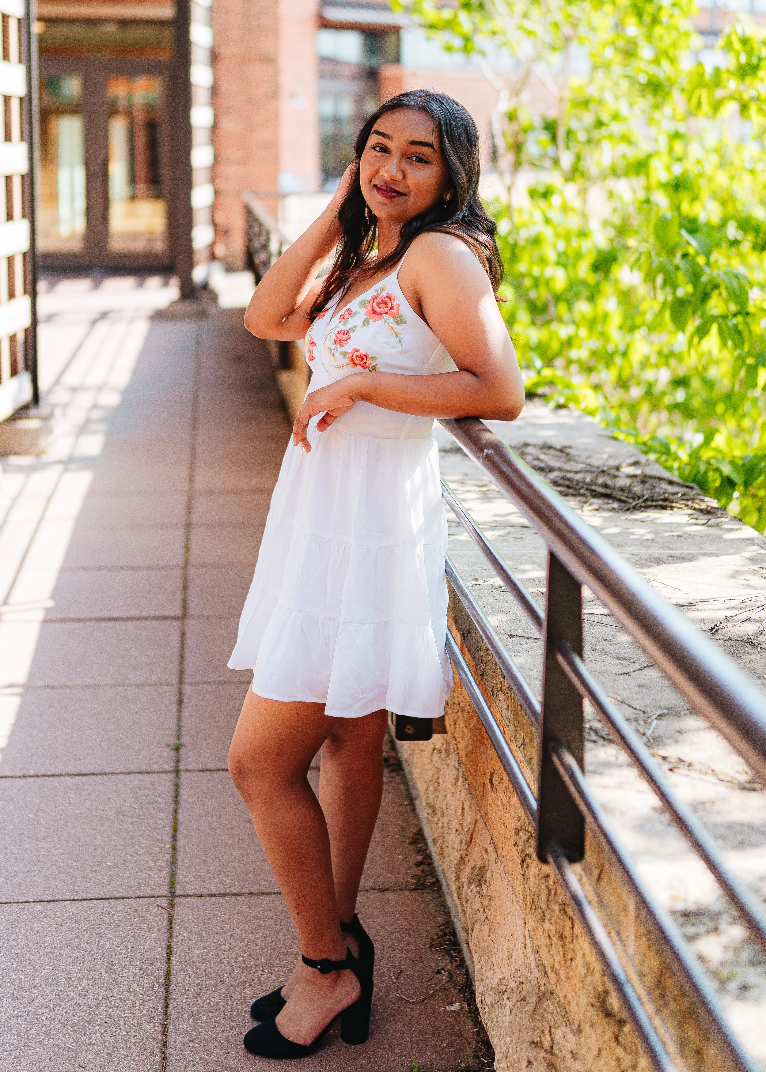 A woman in a white dress with floral embroidery posing outdoors on a sunny day, leaning against a railing on a balcony with greenery and brick buildings in the background.