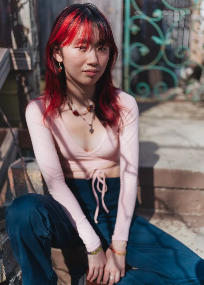 A young woman with red hair and earrings sitting outdoors on steps, wearing a light pink long-sleeve top and dark pants, with jewelry including necklaces and bracelets.