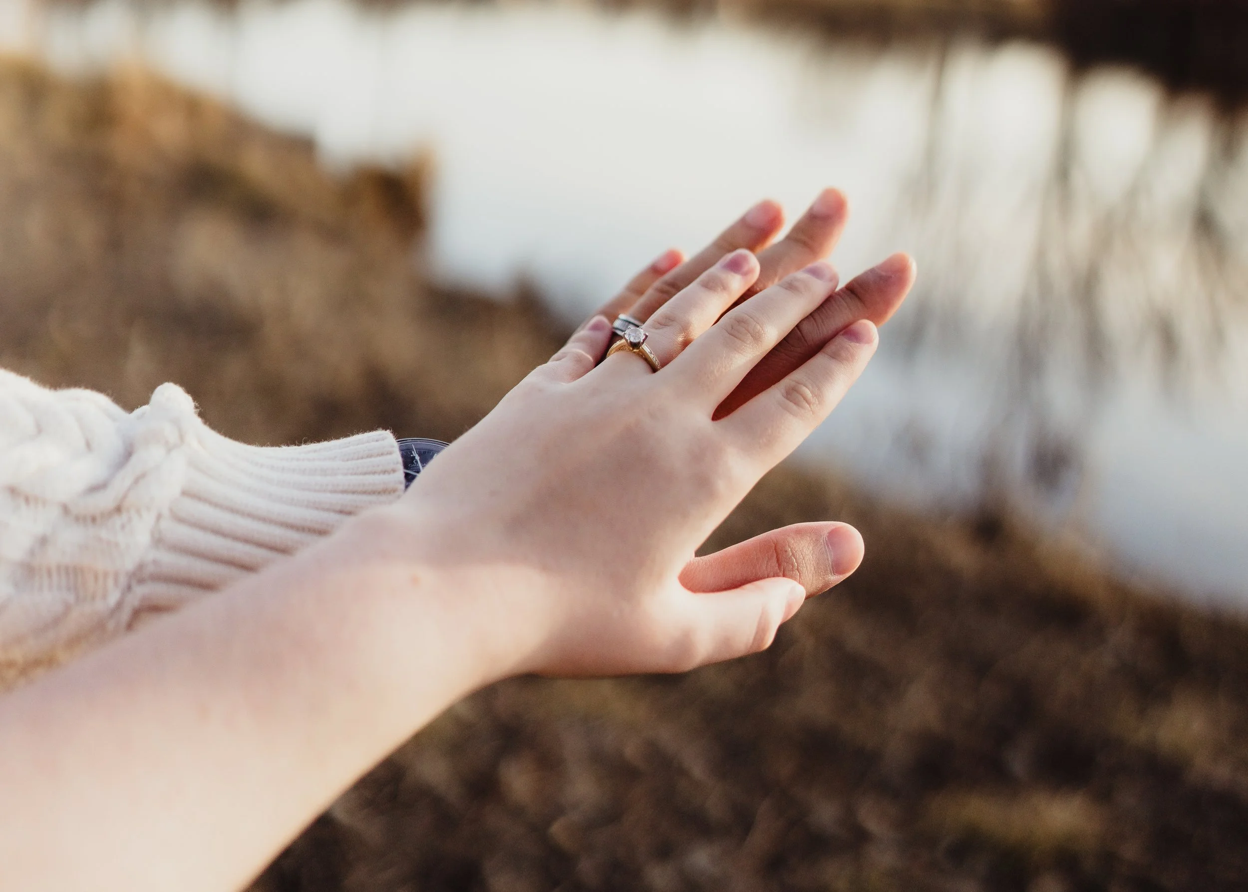 A person's hand with a ring on the finger reaching out toward a blurred natural outdoor background with water and trees.