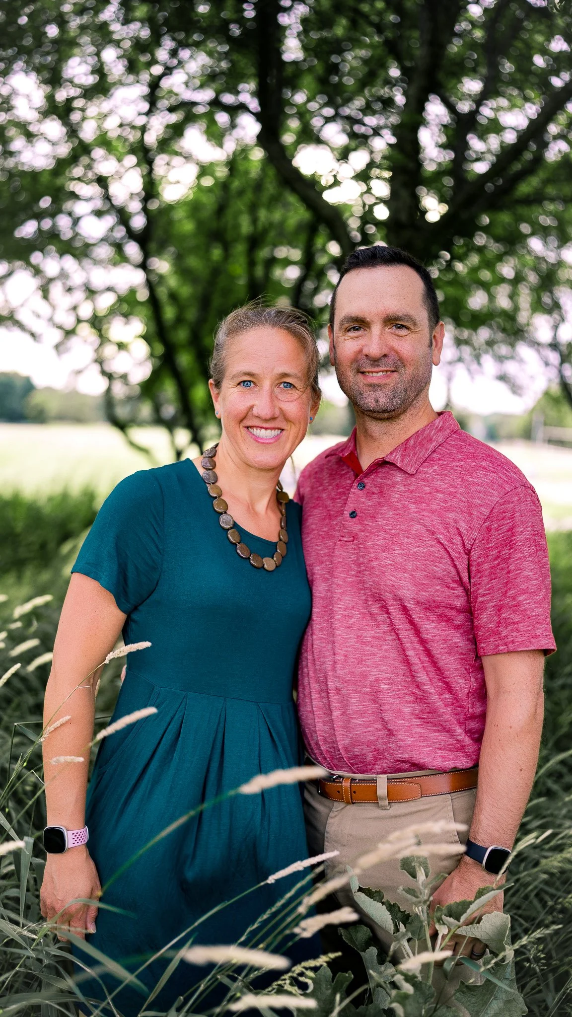A smiling couple standing outdoors near a large tree with green foliage, with a field and sky in the background, during daytime. The woman is wearing a teal dress, a beaded necklace, and a smartwatch, and the man is wearing a red polo shirt and beige