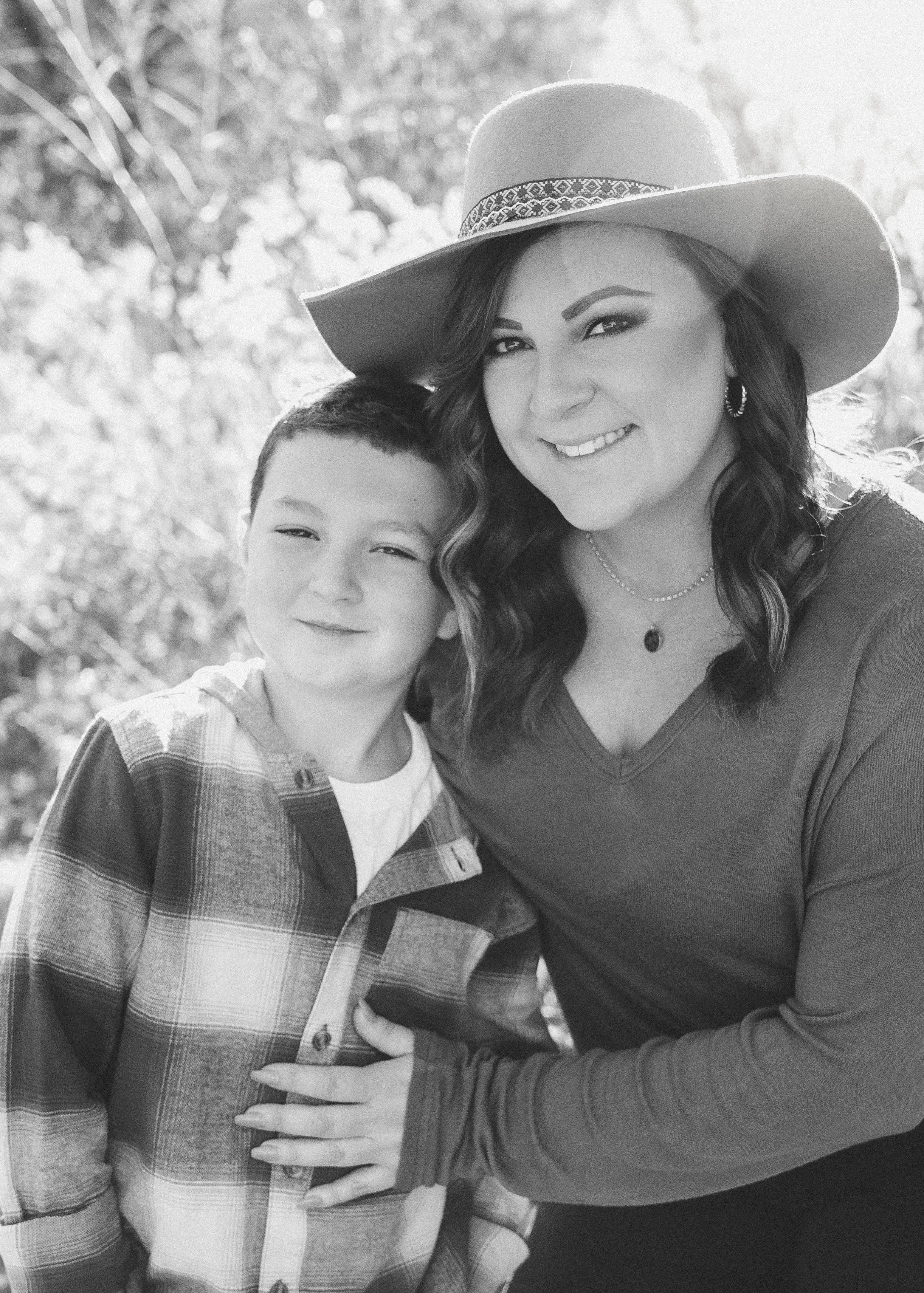 A woman and a young boy smiling outdoors in black and white. The woman is wearing a wide-brimmed hat, earrings, and a necklace. The boy is wearing a plaid shirt.