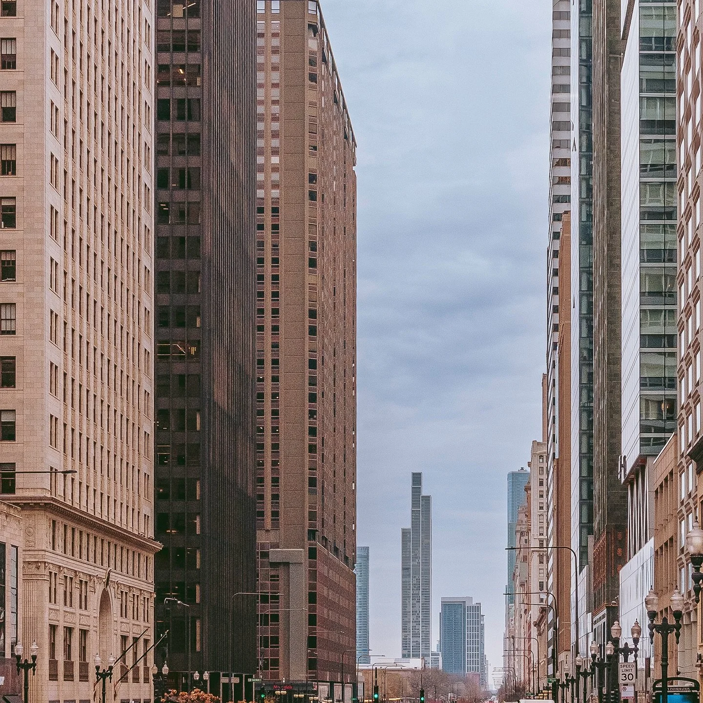 Tall city buildings lining both sides of a street with traffic lights and street lamps, under a cloudy sky, in downtown Chicago.