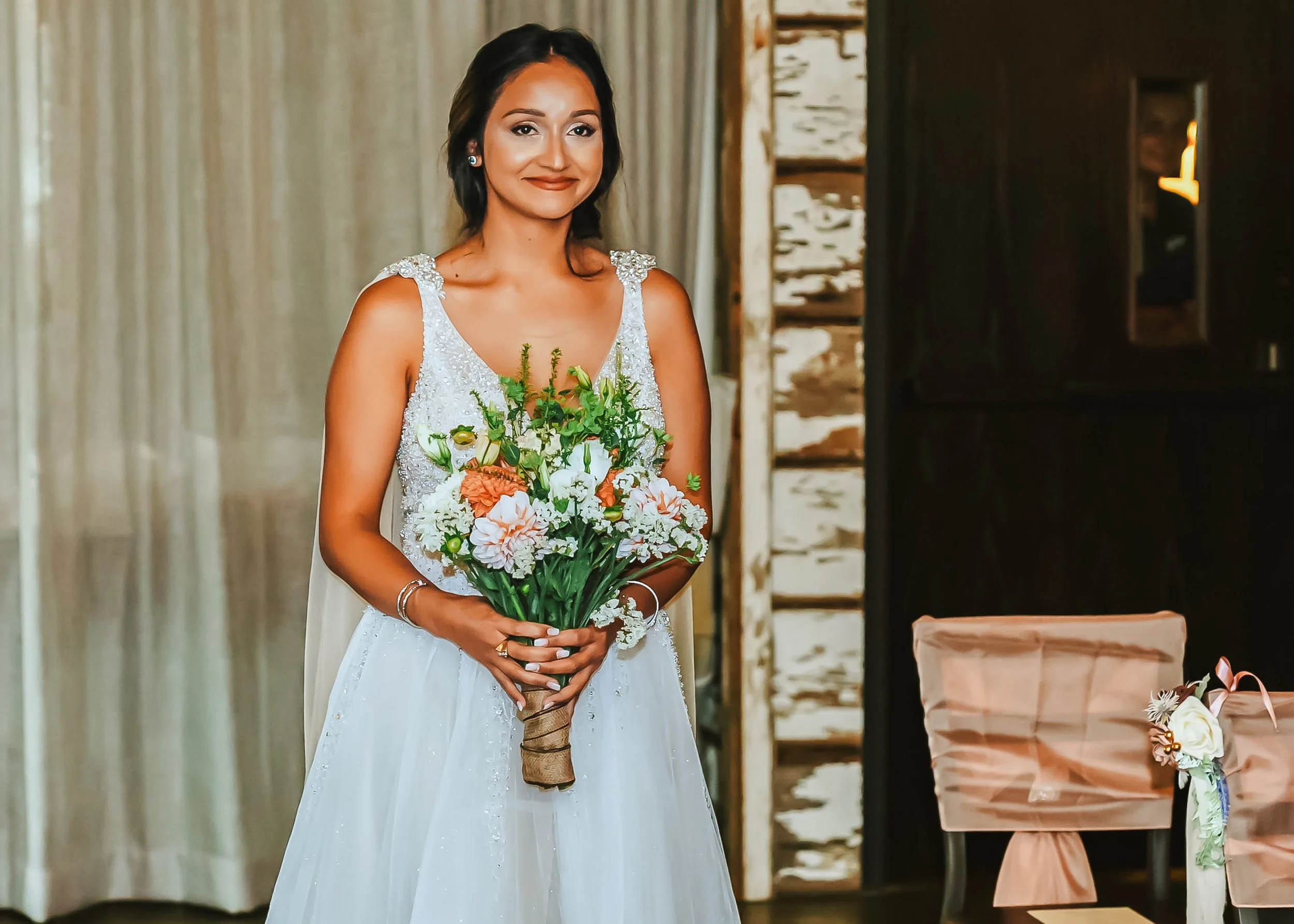 A woman in a white wedding dress holding a bouquet of white and peach flowers inside a rustic venue.