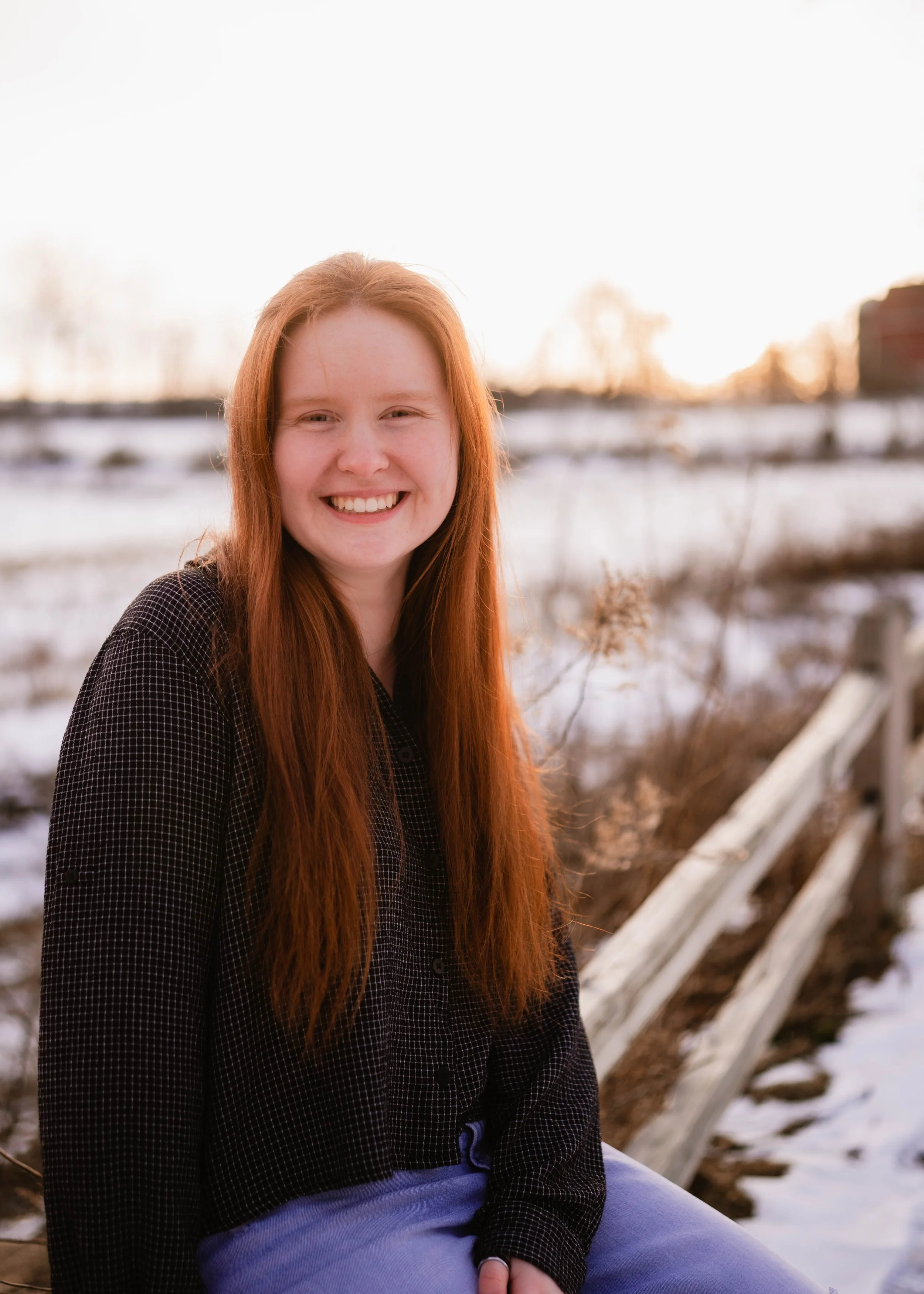 A young woman with long red hair and a black checkered shirt smiling outdoors in a snowy landscape during sunset.