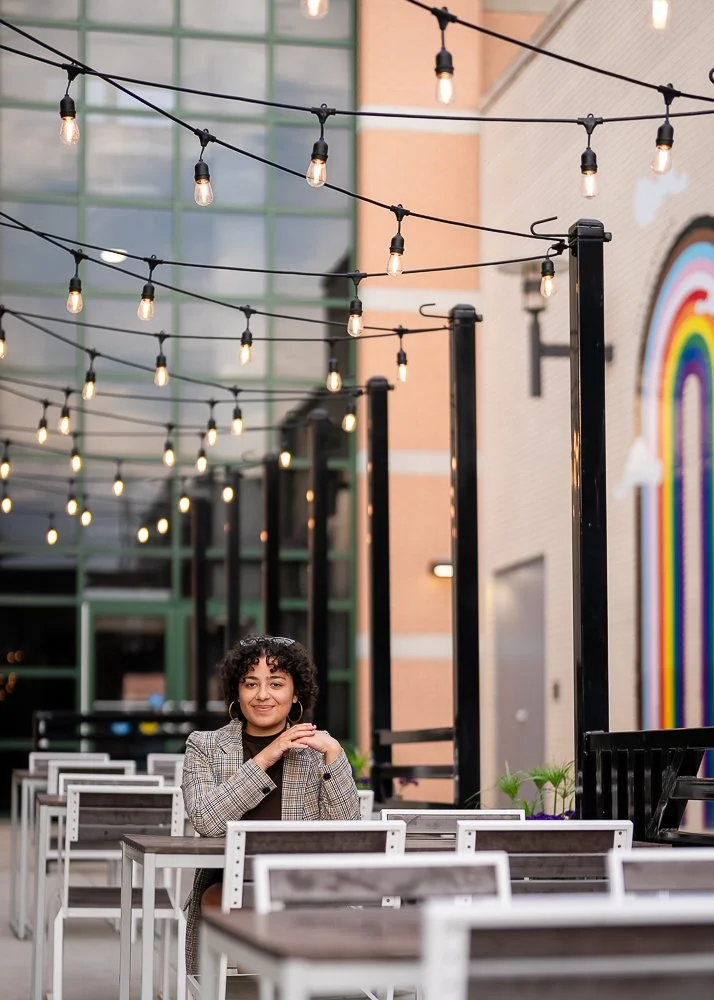 A woman with curly hair and a checkered blazer sitting at an outdoor patio with string lights overhead and empty tables around her, with a colorful rainbow mural on the building wall in the background.