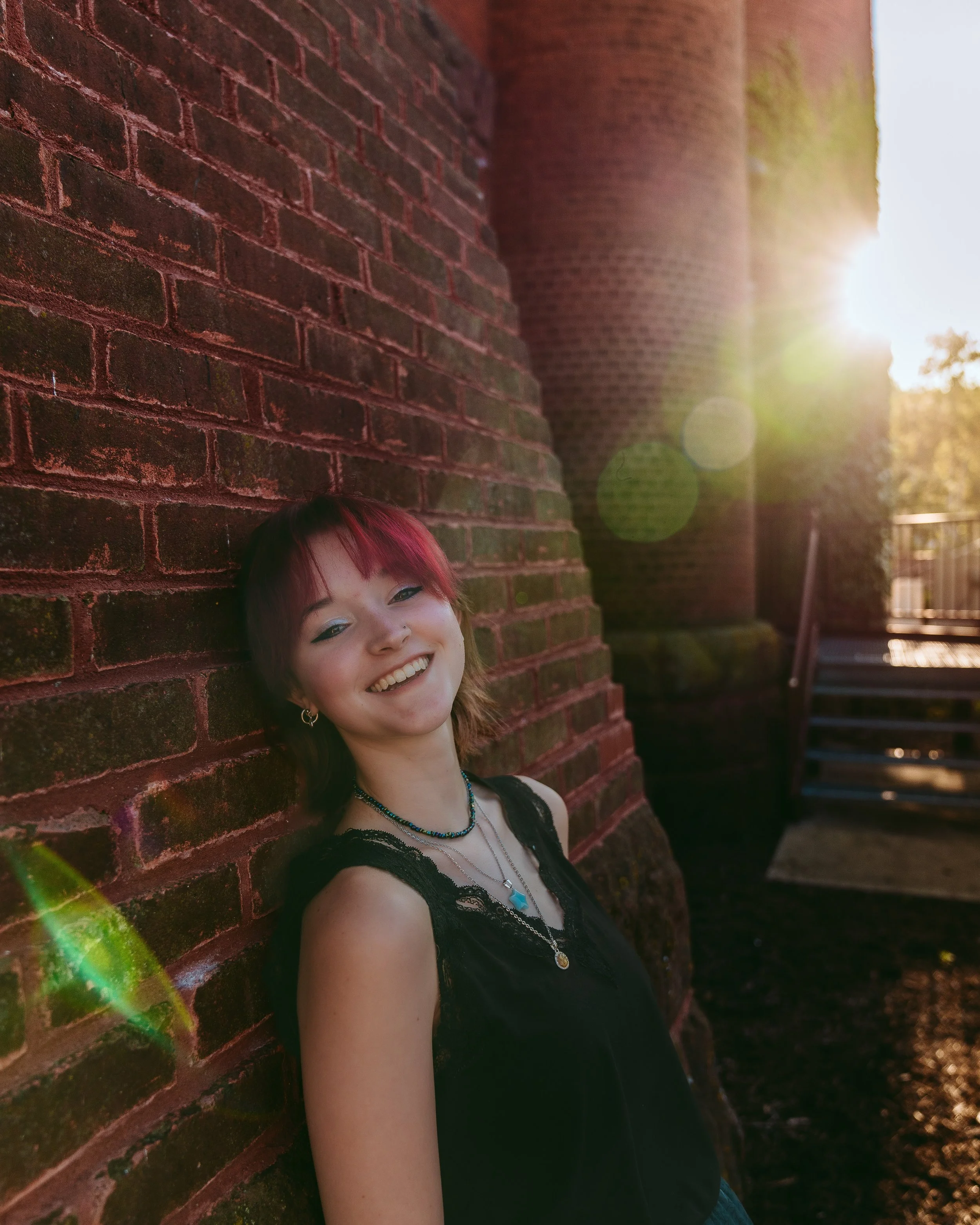 A young woman with short red hair smiling and leaning against a brick wall during sunset.