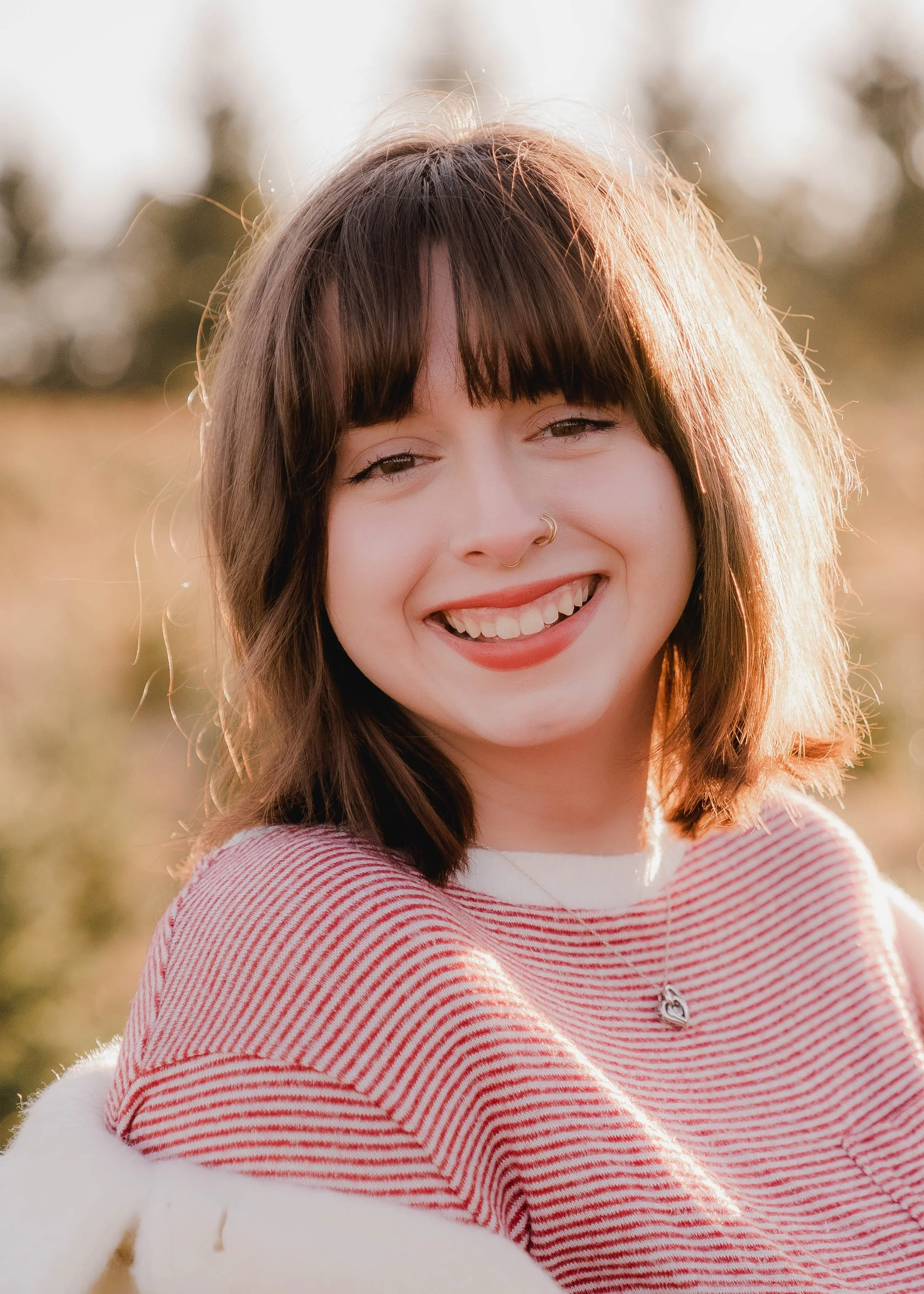 A young woman with dark brown hair and bangs smiling outdoors during golden hour sunlight, wearing a red and white striped shirt and a necklace with a heart pendant.