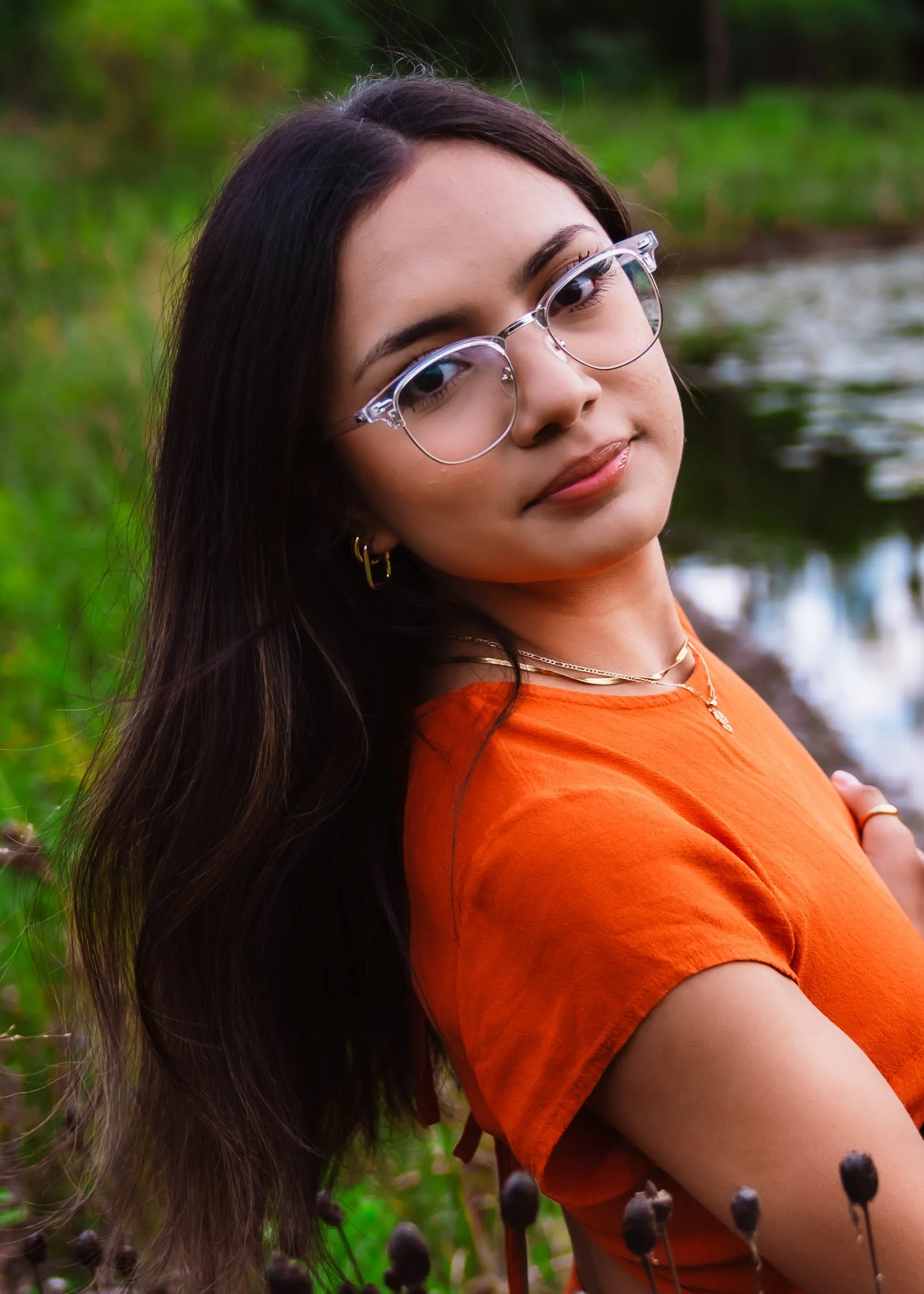 A young woman with long dark hair, wearing glasses and an orange shirt, standing outdoors near a body of water with green trees in the background.