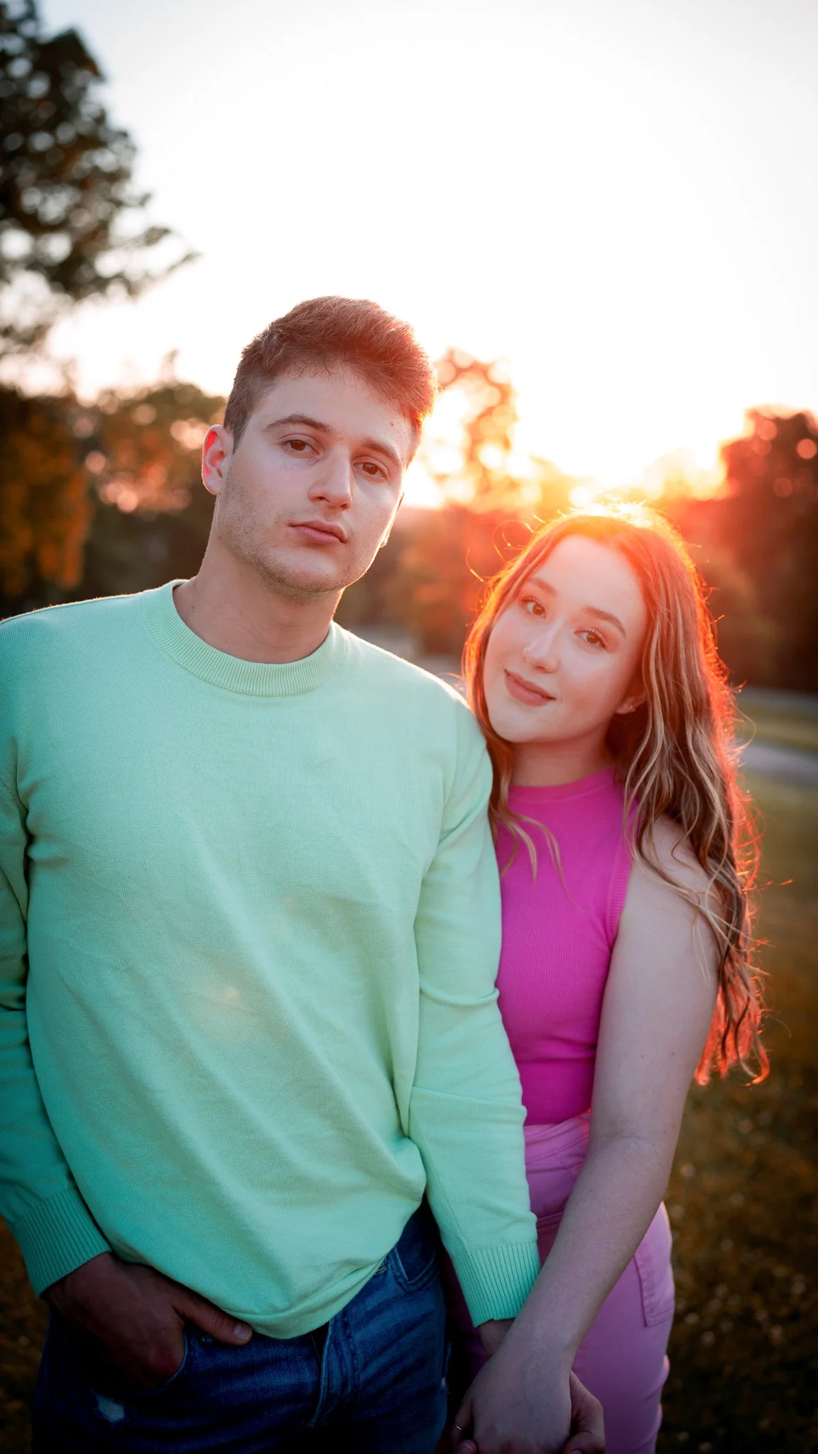 A young man and woman stand close outdoors during sunset, with trees in the background. The man wears a light green sweater and has short dark hair. The woman wears a pink top and has long wavy hair. They look towards the camera, with the sunset cast