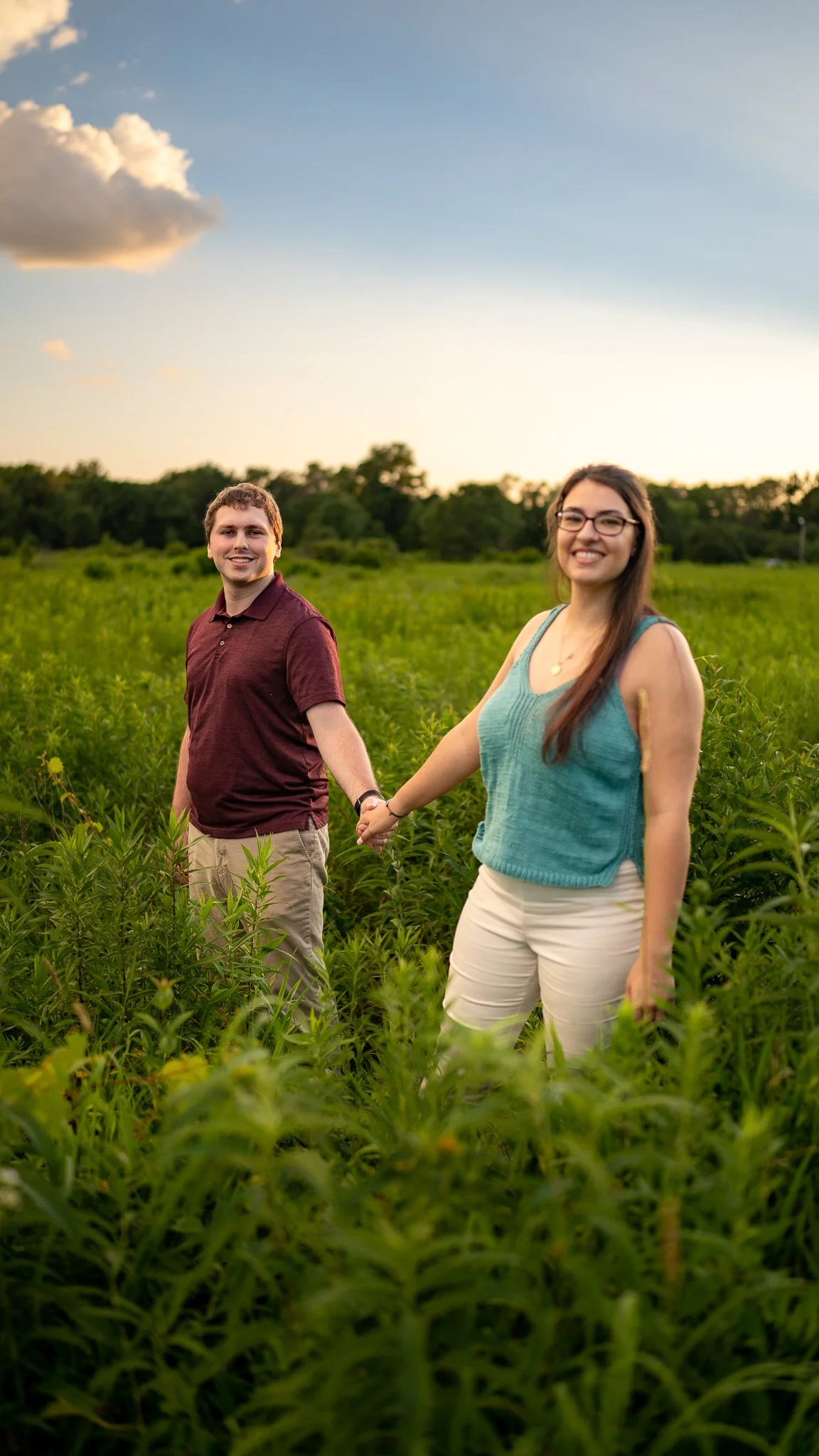A smiling couple holding hands in a field of tall green plants, with a sunset sky and clouds overhead.