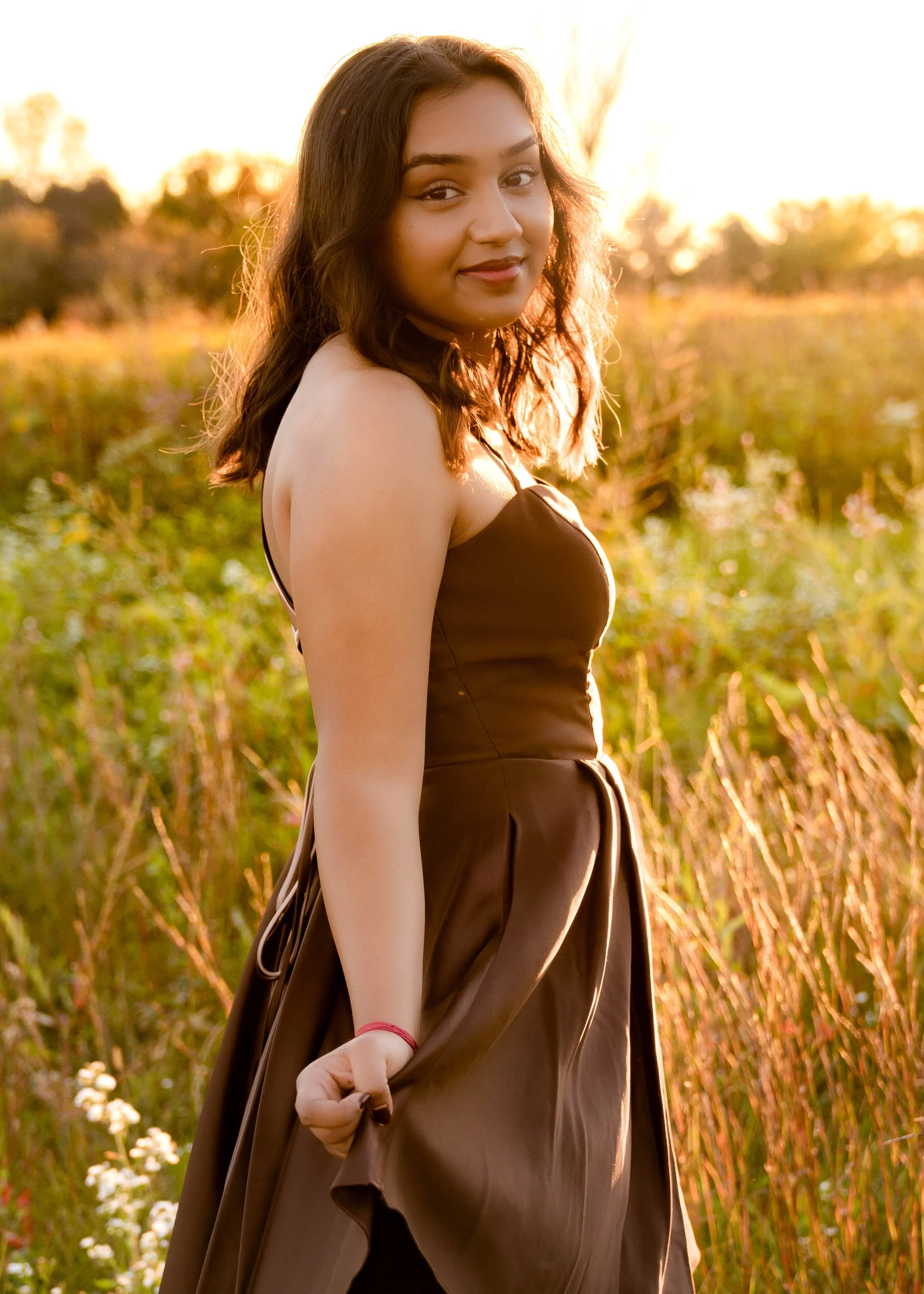 A woman with dark hair in a brown dress standing outdoors in a field during sunset.