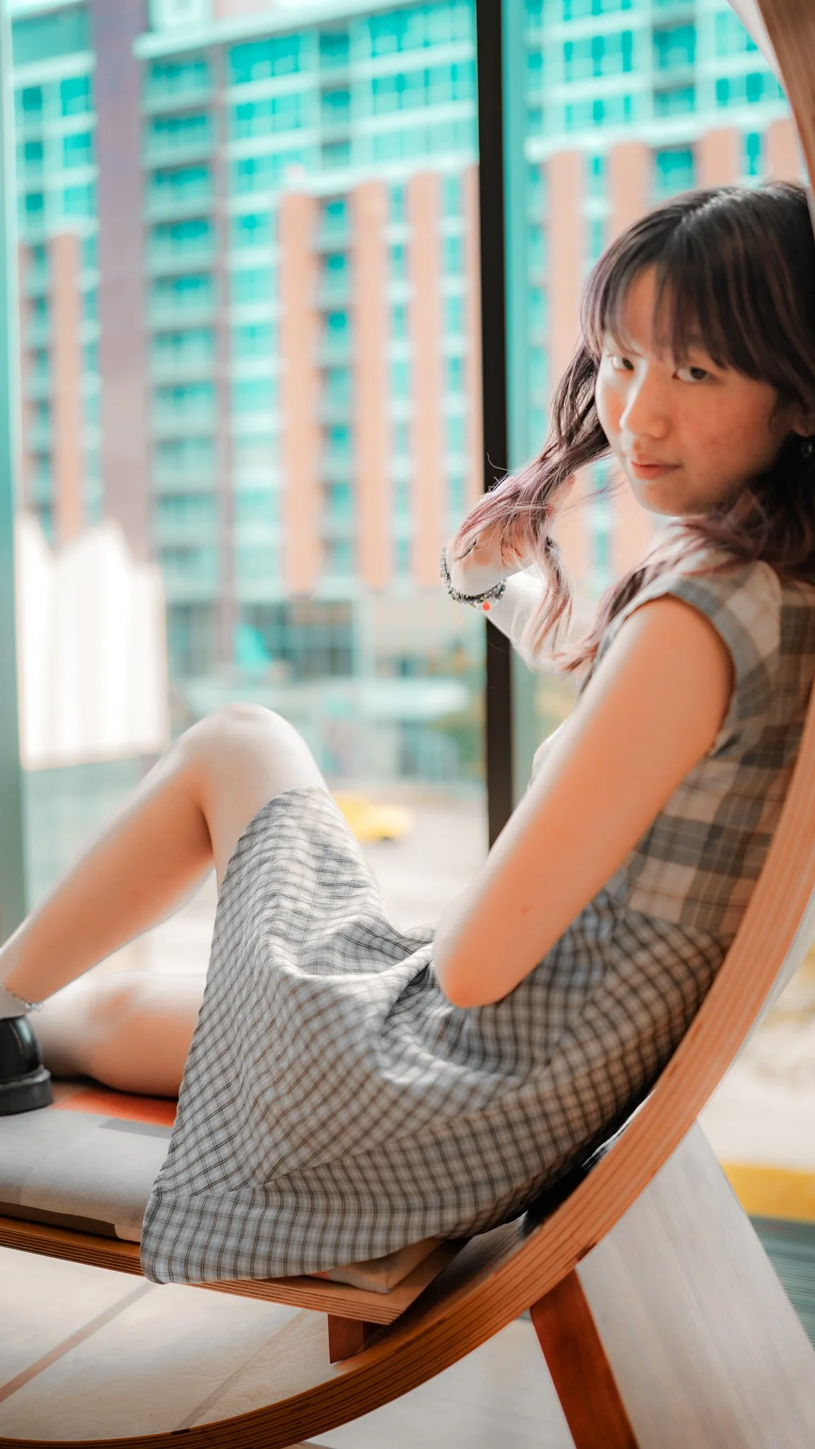 A young girl with wavy hair sitting on a wooden lounge chair near a large window, with a cityscape of tall buildings visible outside.