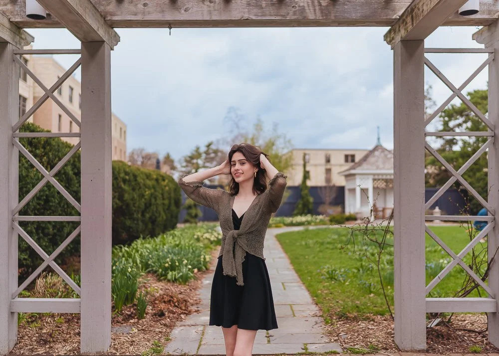 A young woman with dark hair in a green cardigan over a black dress, standing under a wooden archway in a garden, smiling with her hands in her hair.