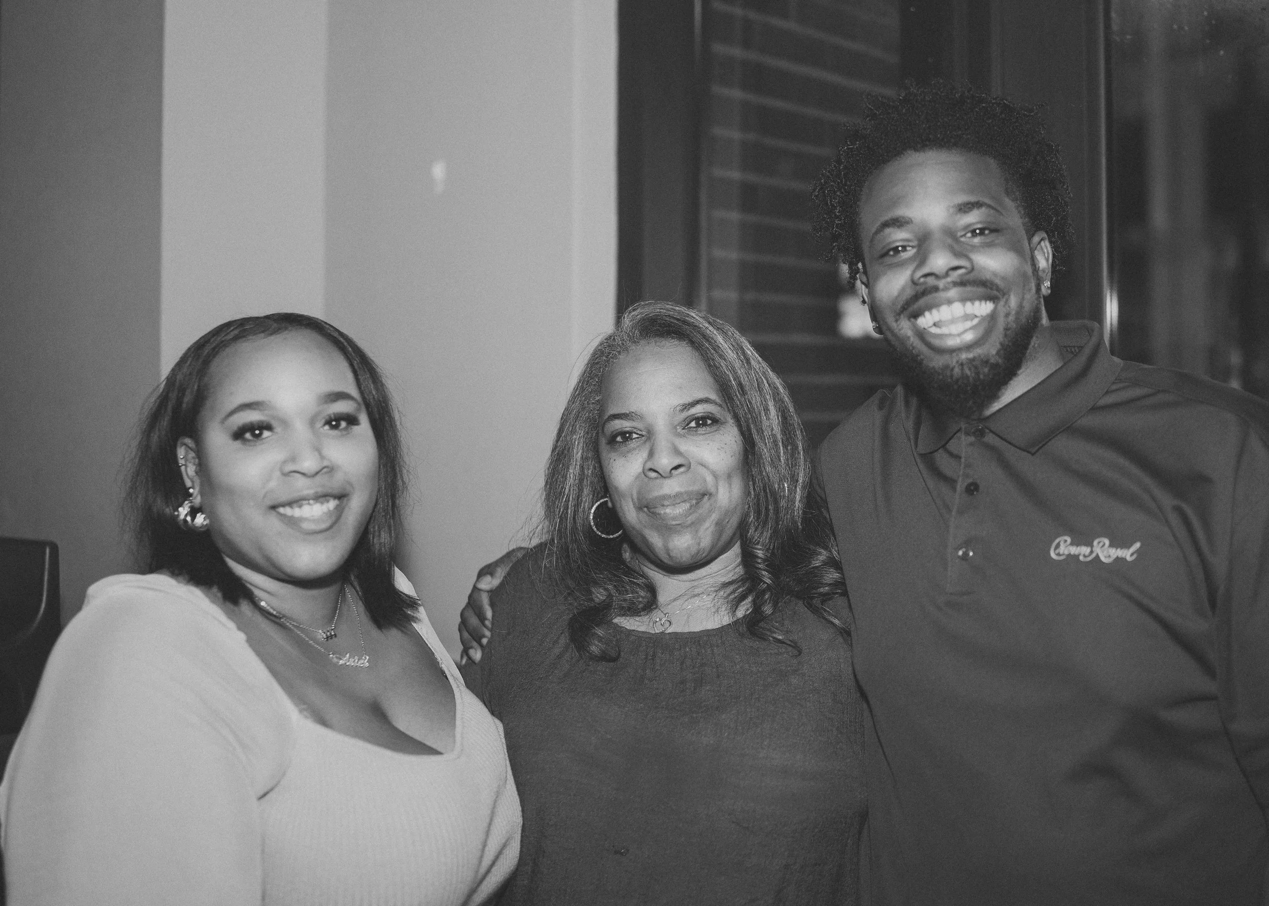 Three people standing close together and smiling in a black-and-white photo. The woman on the left has shoulder-length hair, earrings, and a light-colored top. The woman in the middle has shoulder-length curly hair, hoop earrings, and a dark top. The