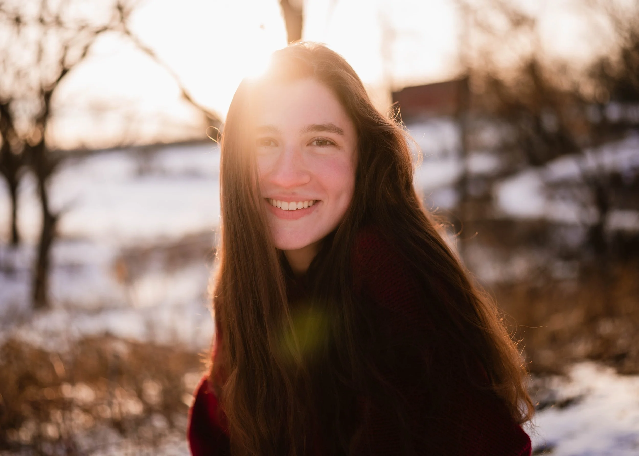 A young woman with long brown hair smiling outdoors in a winter setting with a snow-covered landscape and trees in the background, backlit by sunlight.