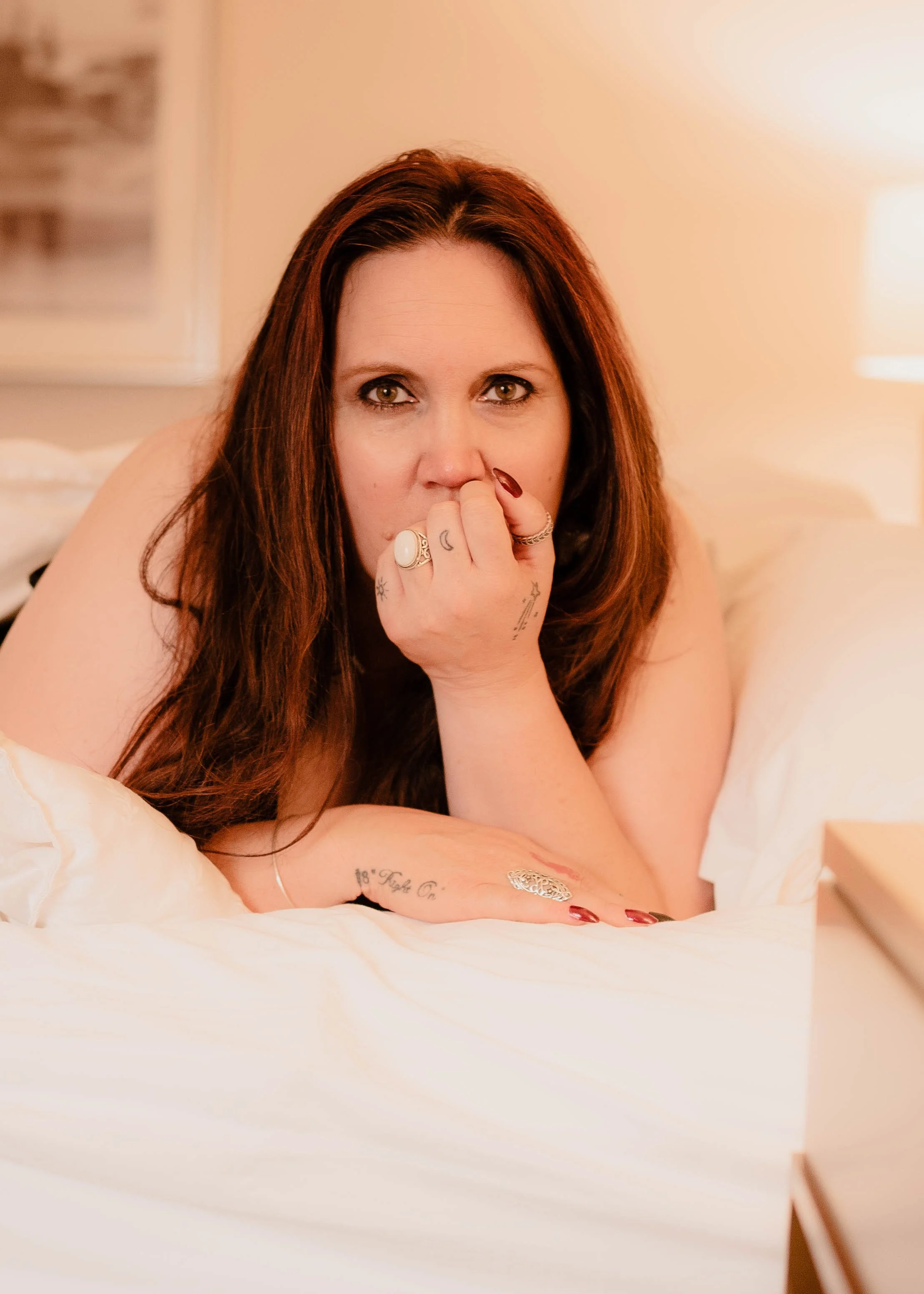 A woman with long reddish-brown hair lying on a bed, resting her head on her hand with tattoos and rings, looking directly at the camera in a warmly lit room.