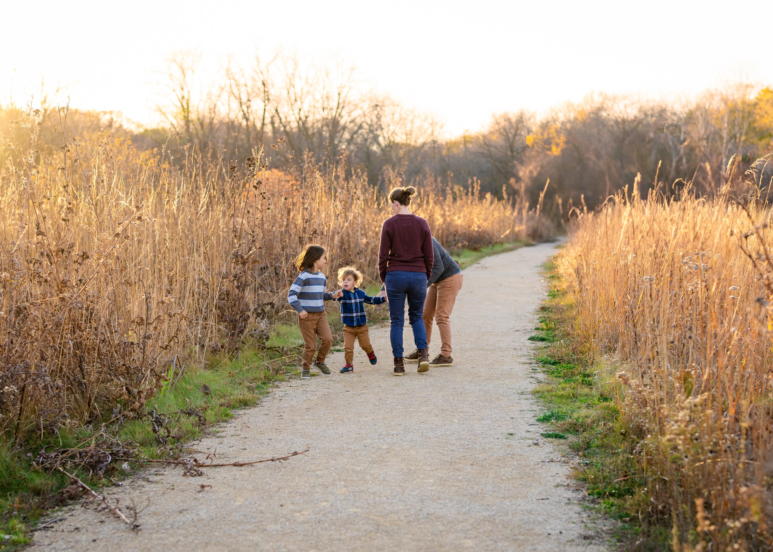 Family walking on a dirt path through a field of tall, brown grass during sunset, with bare trees in the background.