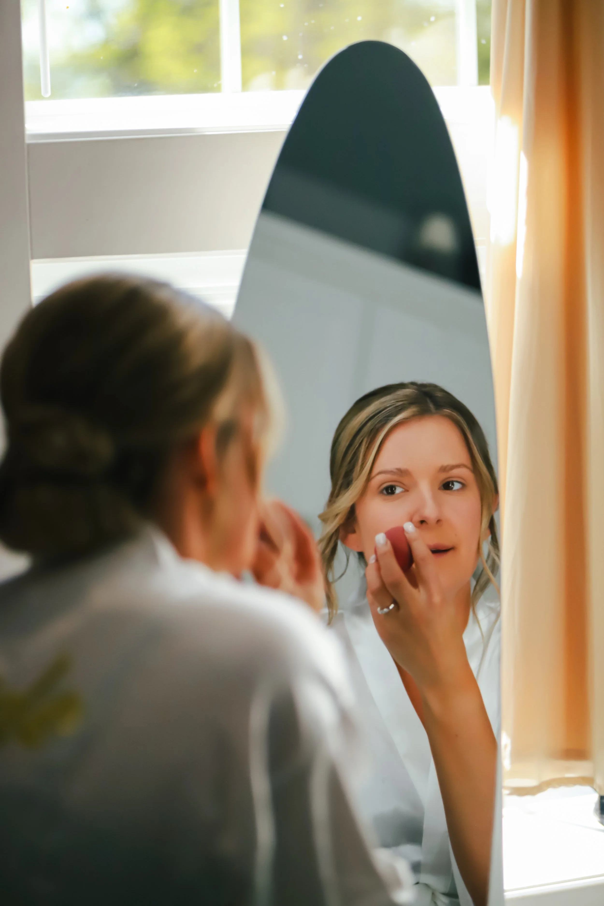 A woman applying makeup in front of a mirror, with sunlight coming through a window in the background.