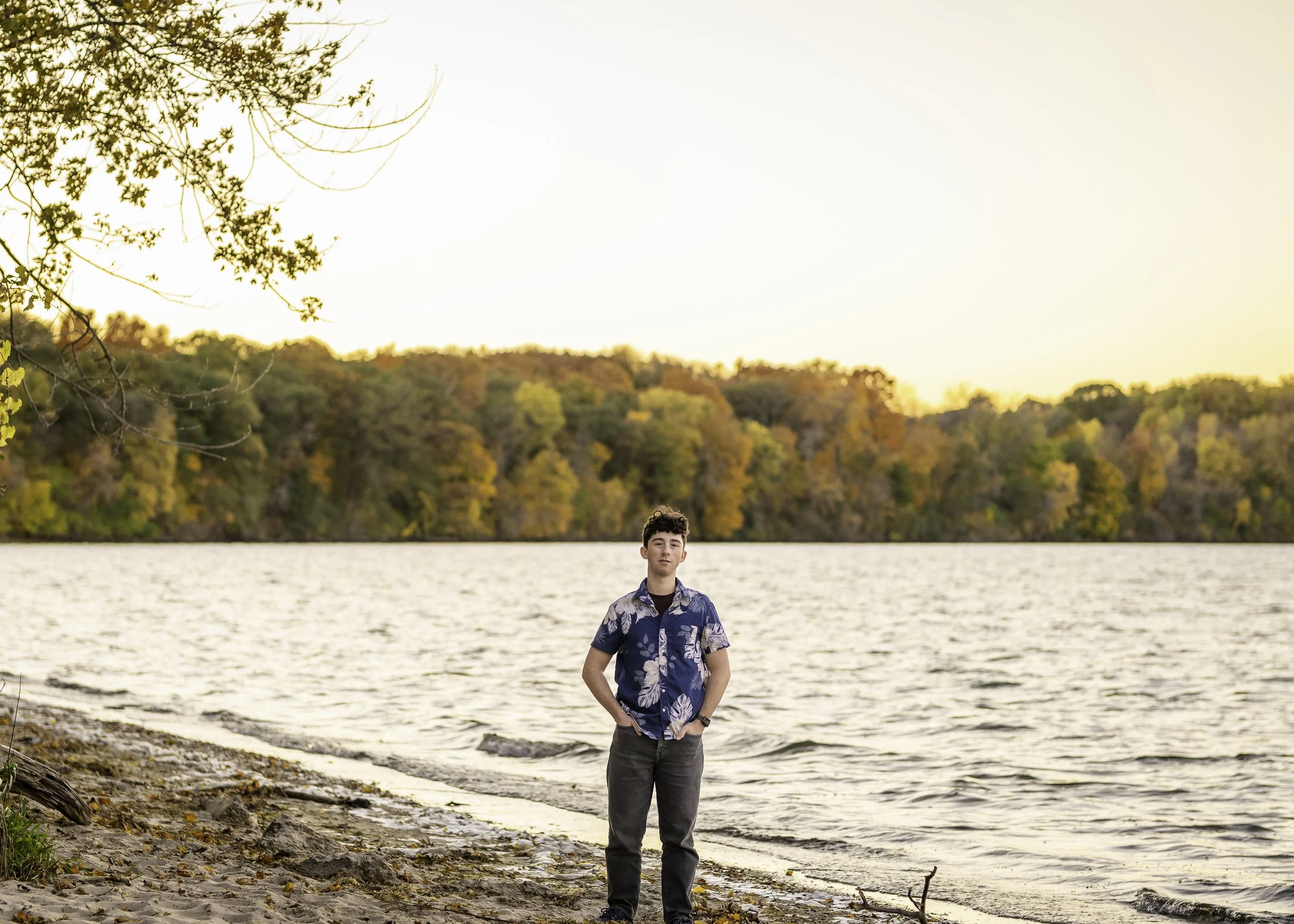 A young man standing on a sandy shore by a lake during sunset, with trees in autumn colors in the background.