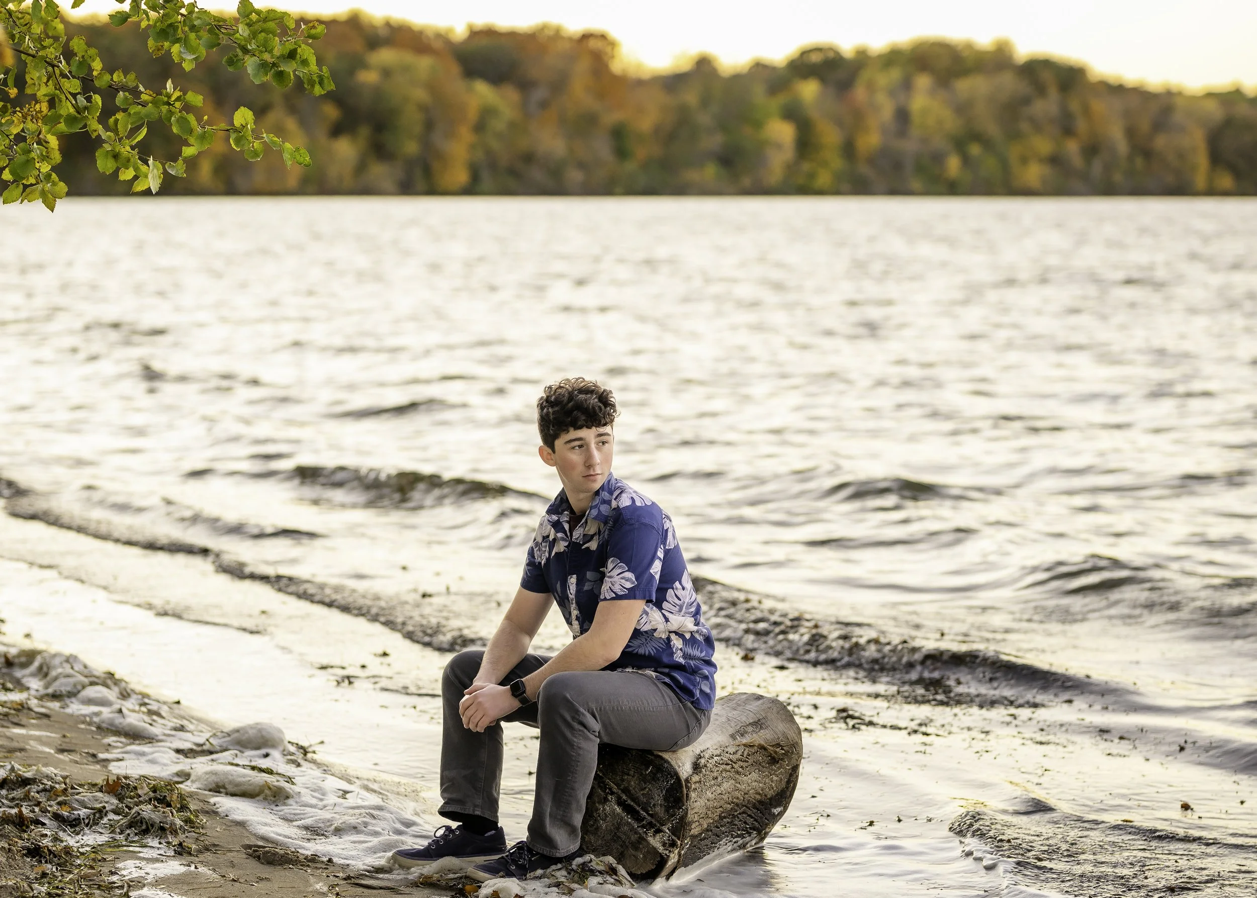 A young man sitting on a log at the edge of a lake during fall, wearing a blue Hawaiian shirt and gray pants, with autumn trees in the background.
