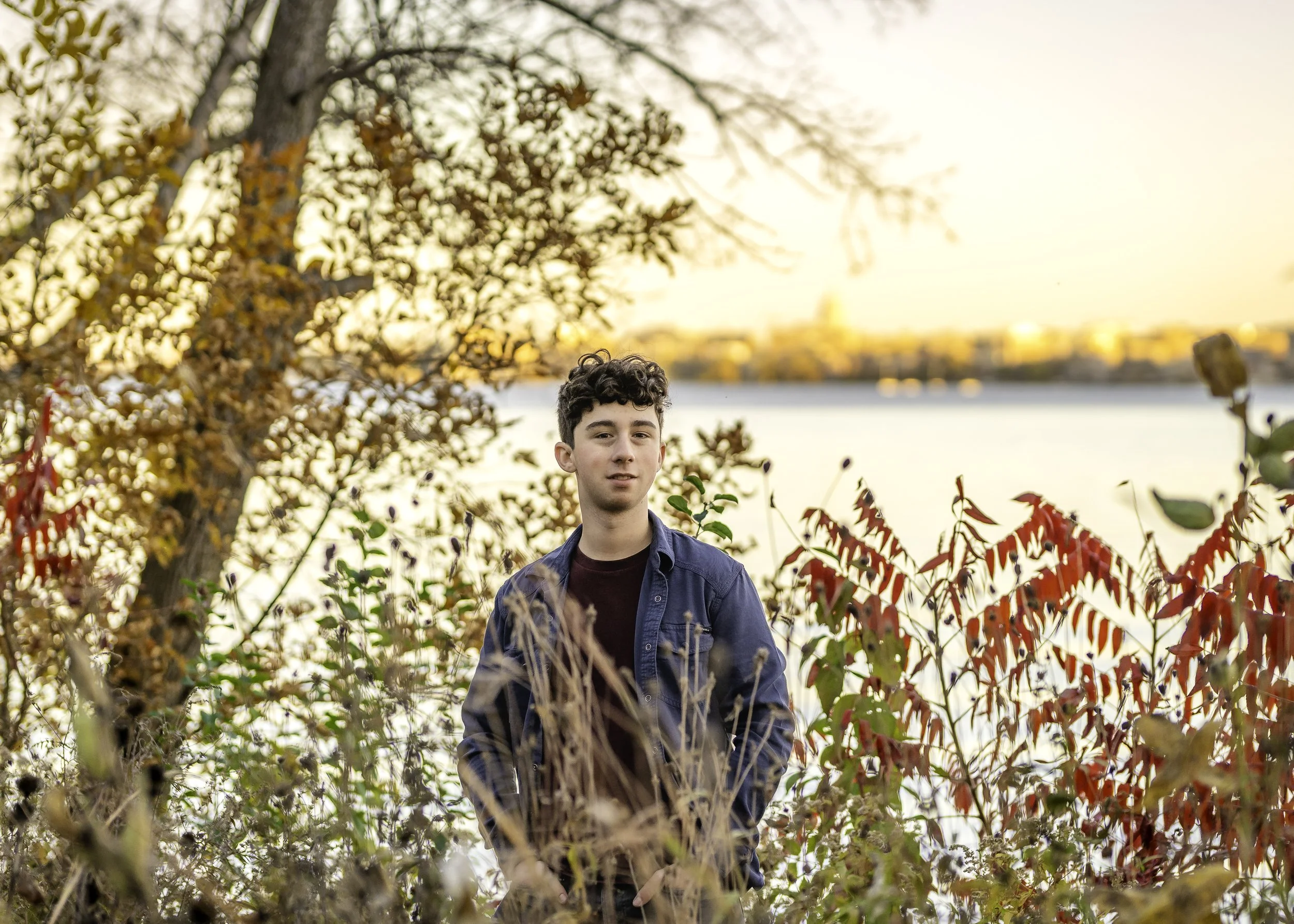 A young man standing outdoors among fall foliage near a body of water, with city buildings in the distance at sunset.