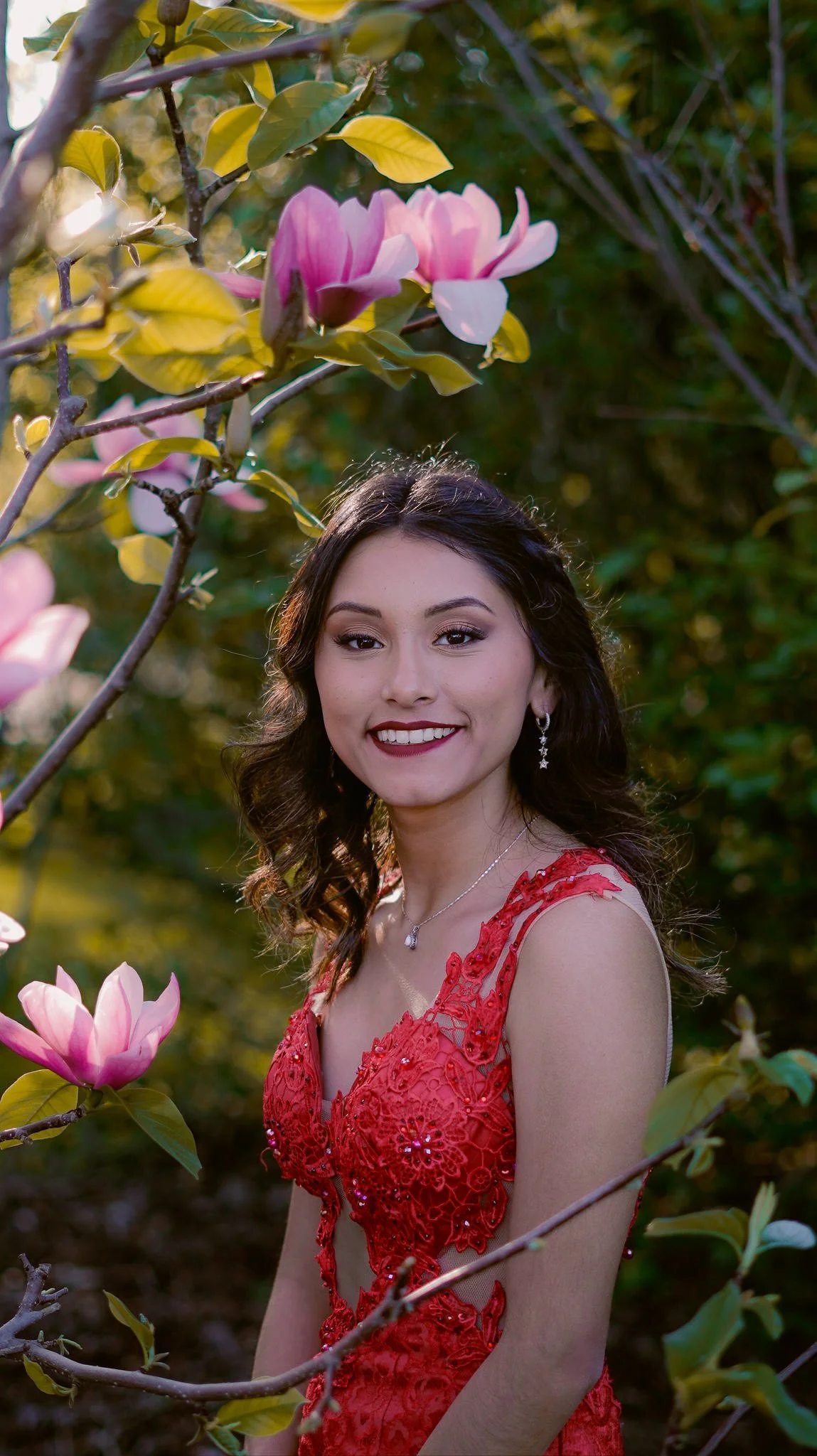 A young woman in a red dress standing among blooming pink magnolia flowers, smiling at the camera.