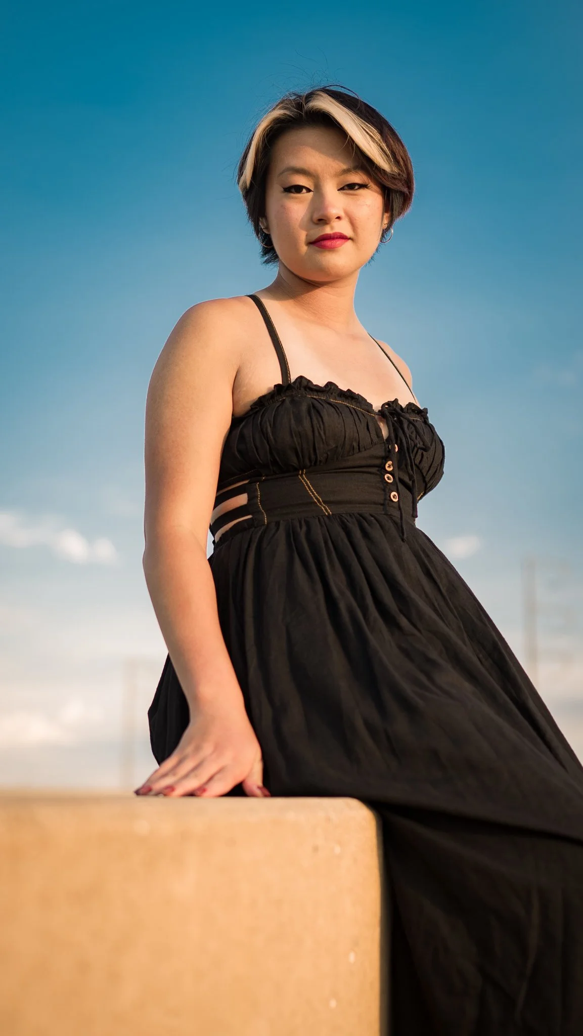 A woman in a black dress looking at the camera, standing outdoors against a blue sky with some clouds.
